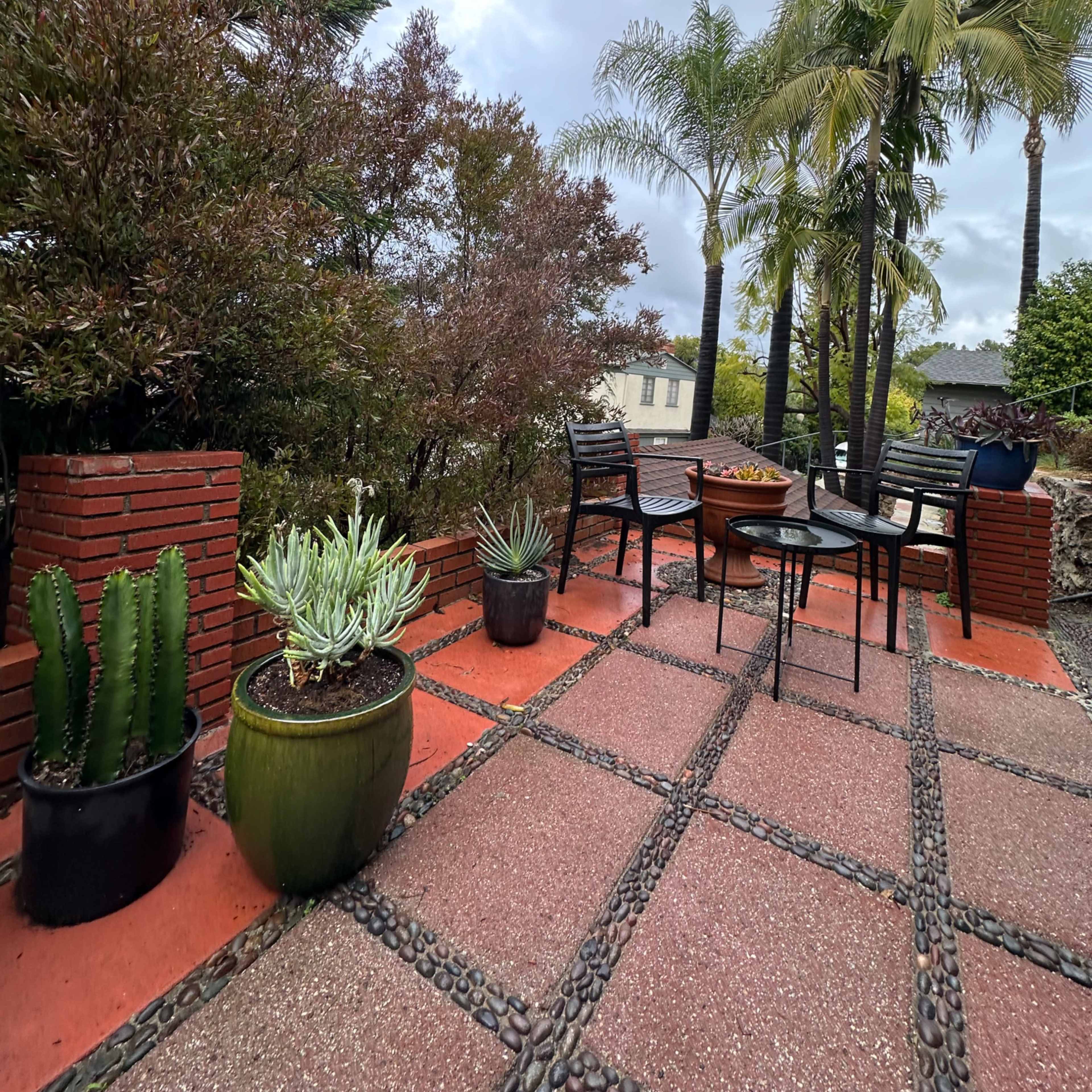 A patio area features brick walls, potted plants, and a small table set with chairs under a cloudy sky.