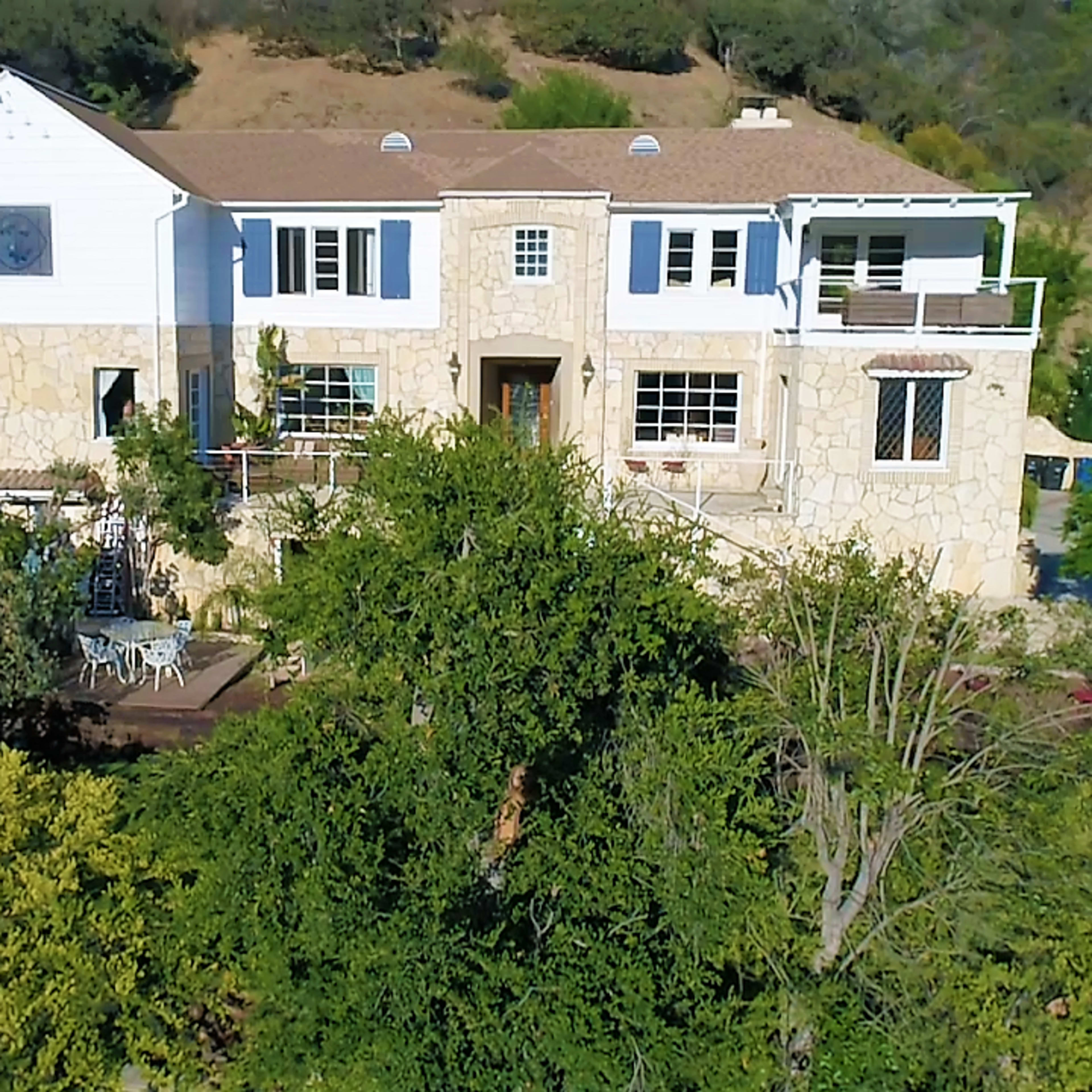 A two-story stone and white house surrounded by greenery on a hillside.