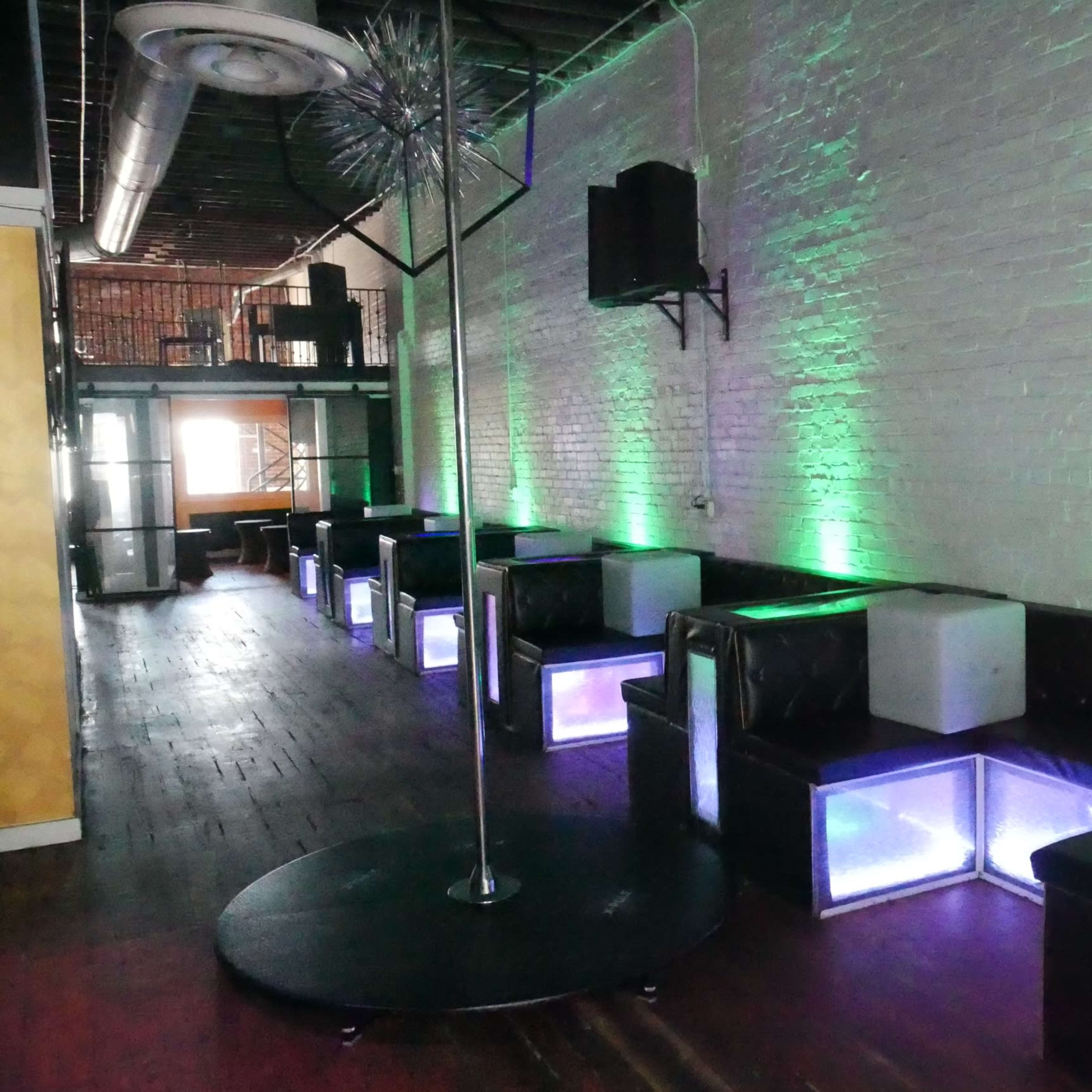 The interior of a nightclub features a row of illuminated seating areas and a central dance pole, with green lighting highlighting the space.