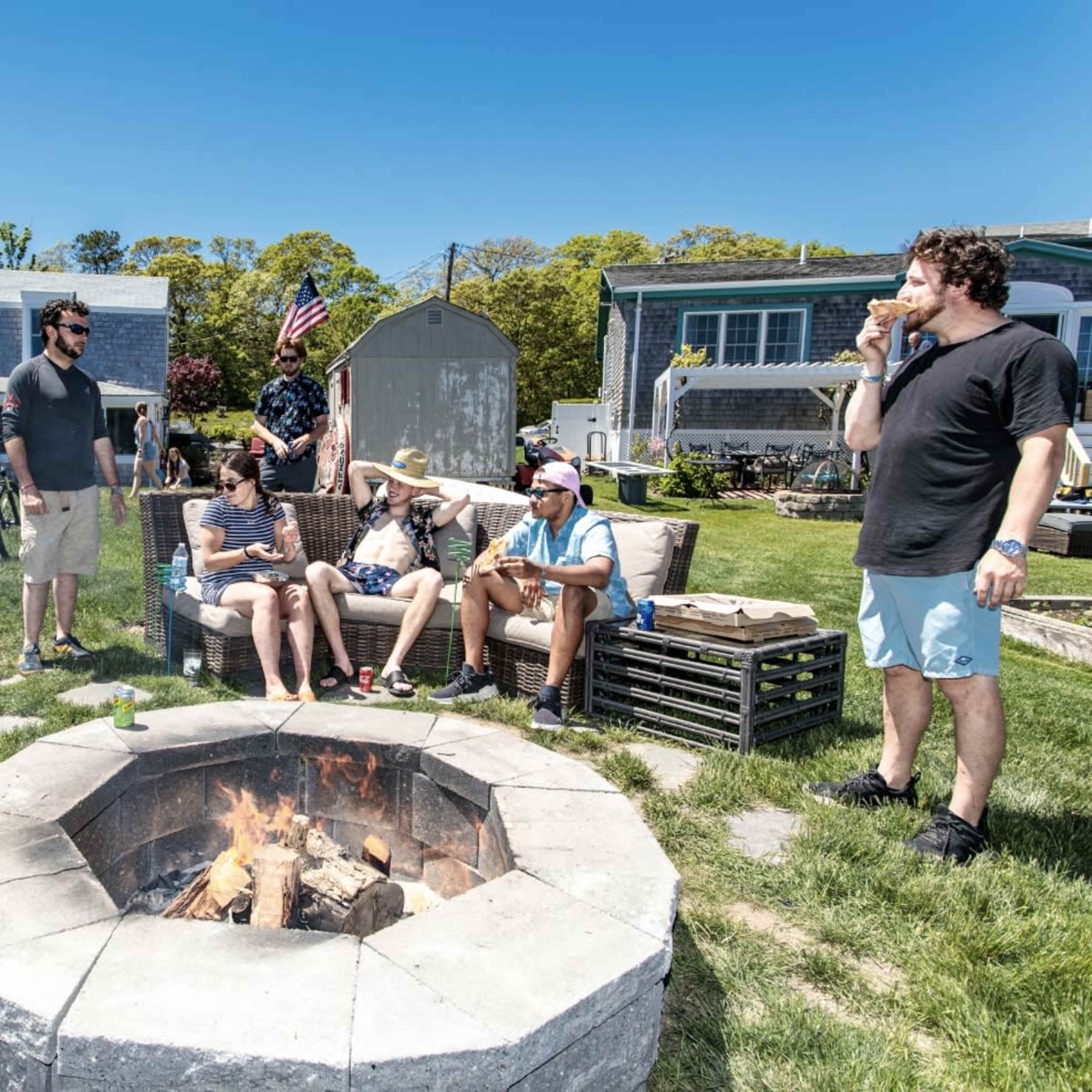 A group of six people gathers around a fire pit in a grassy area, with bicycles nearby and a house in the background.