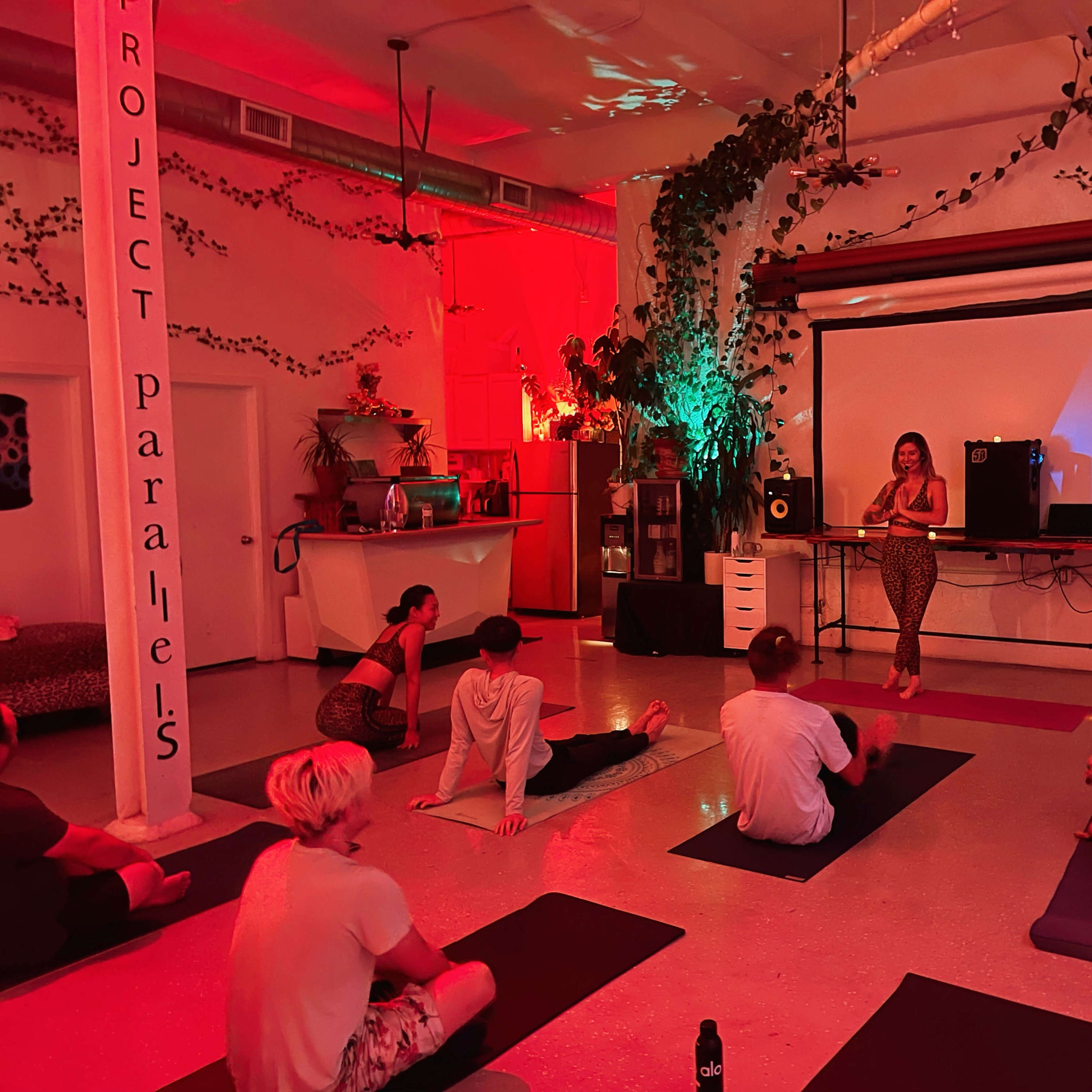 A fitness instructor leads a group yoga session in a dimly lit studio with vibrant red and green lighting.