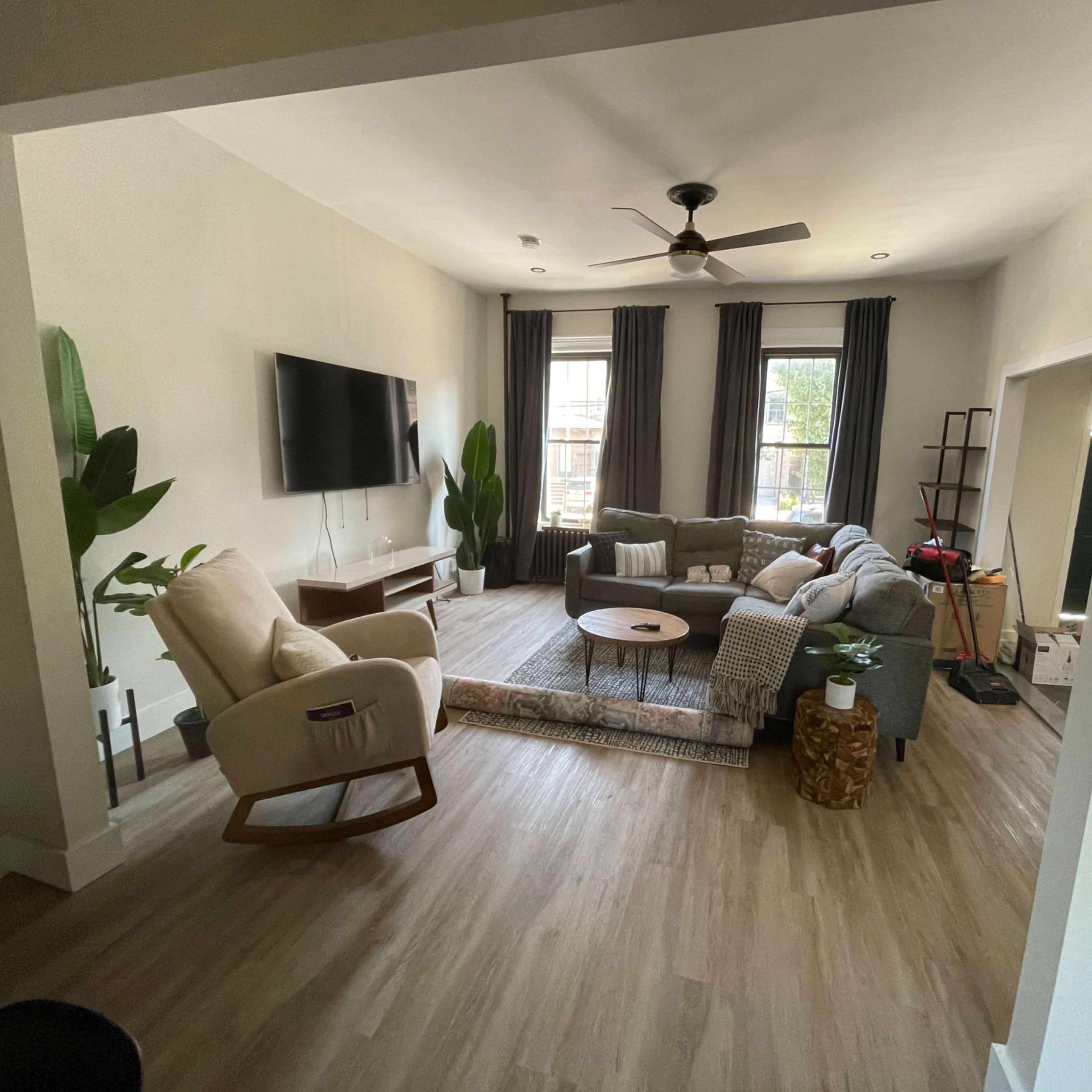 A living room with a gray sectional sofa, a round coffee table, a television mounted on the wall, and large windows with curtains.