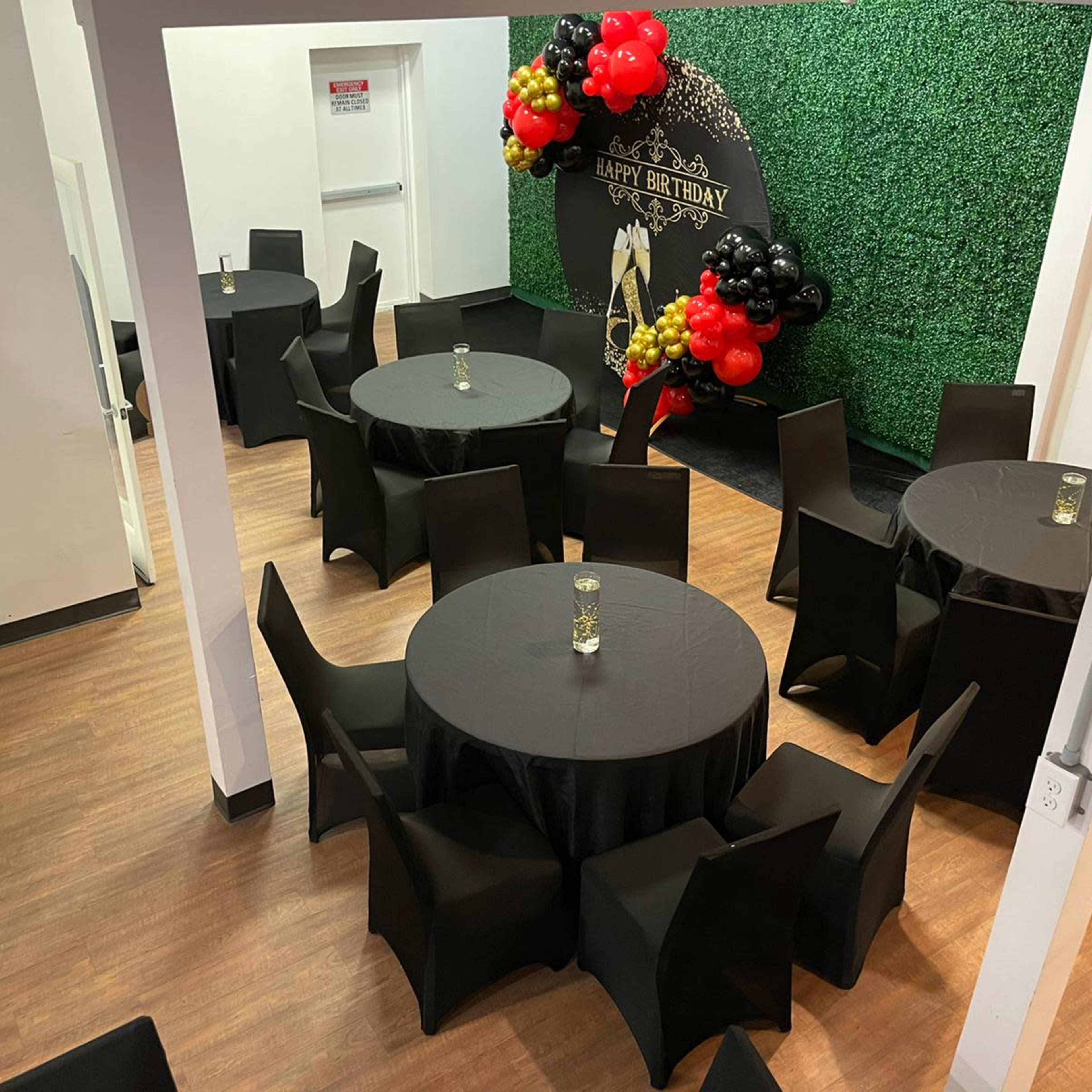 A decorated event space with round tables covered in yellow tablecloths and a large floral backdrop featuring the letters "MRS."