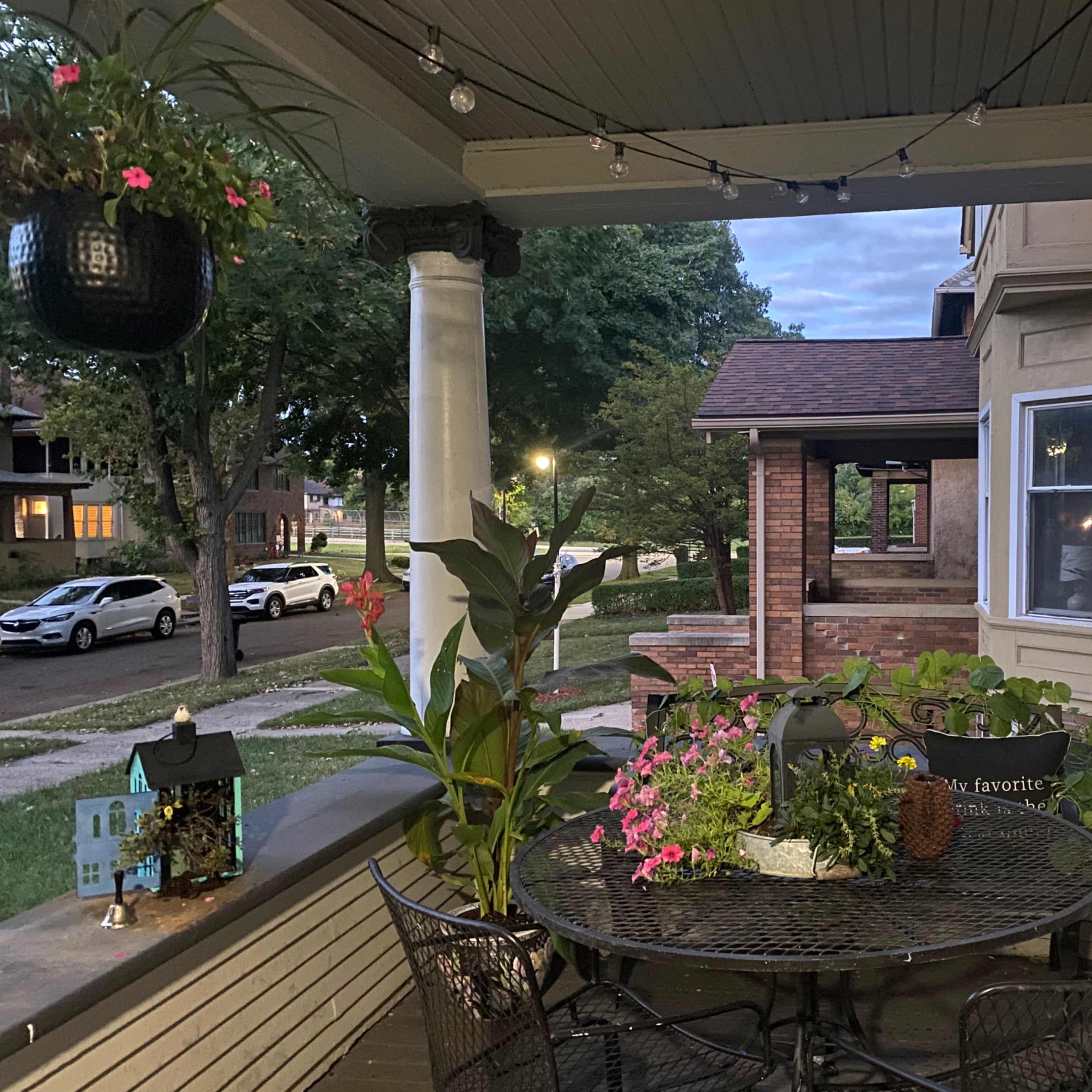 The scene shows a porch with a round table surrounded by plants and flowers, illuminated by string lights, overlooking a neighborhood street with parked cars.