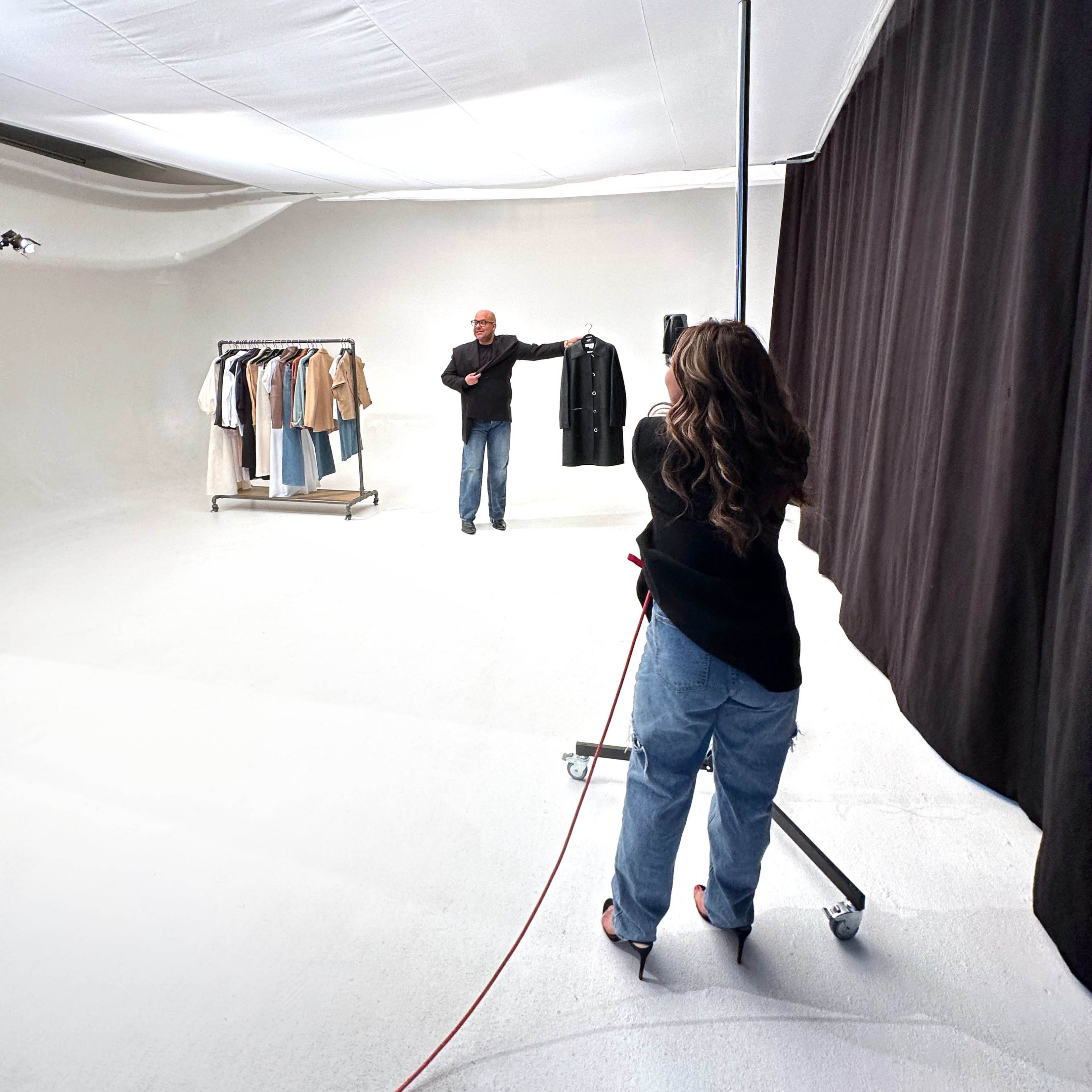 A person stands in a studio holding a coat while another person photographs them from a distance.