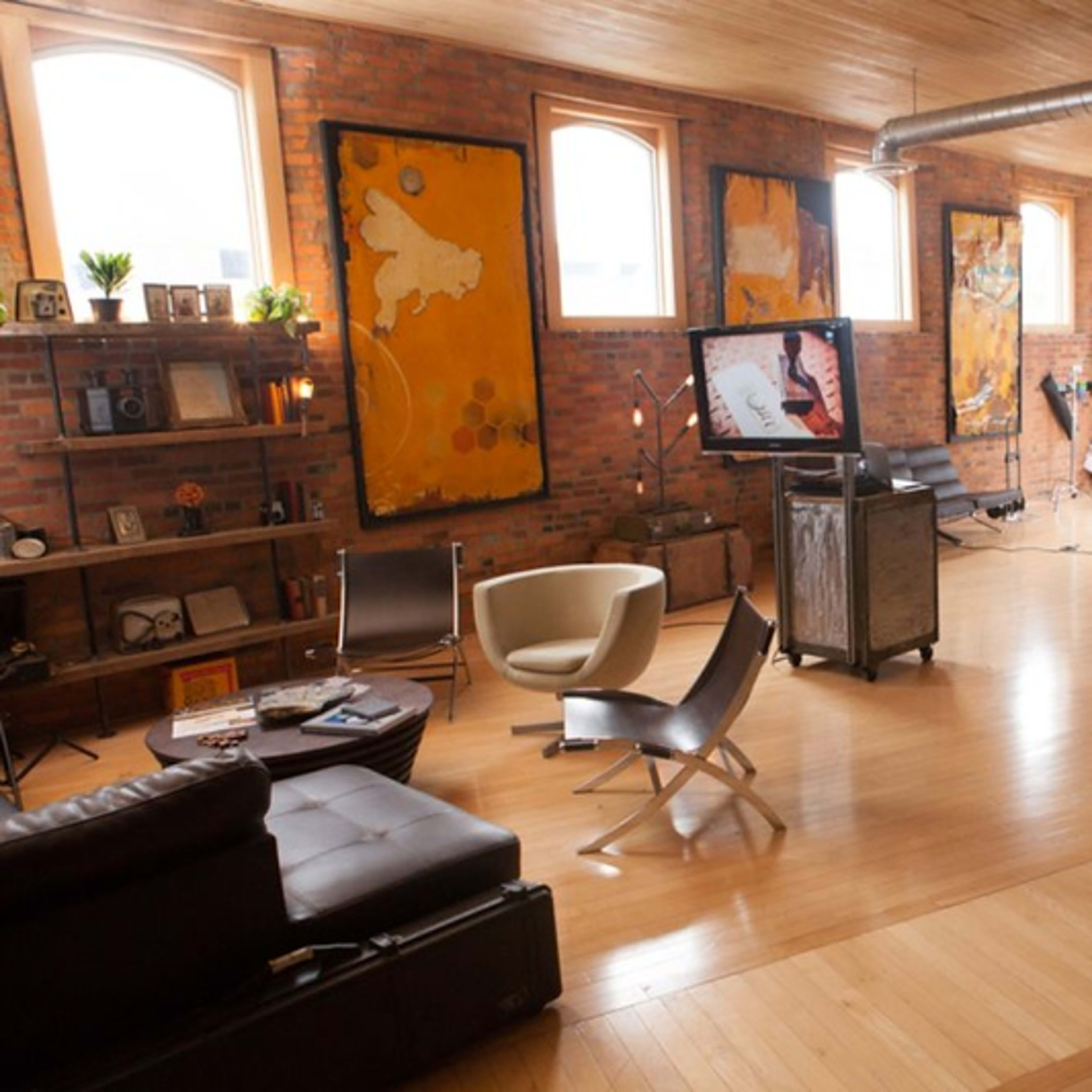 A spacious studio with wooden floors, large windows, and various pieces of furniture, including modern chairs and a leather couch, along with photography equipment set up in one corner.