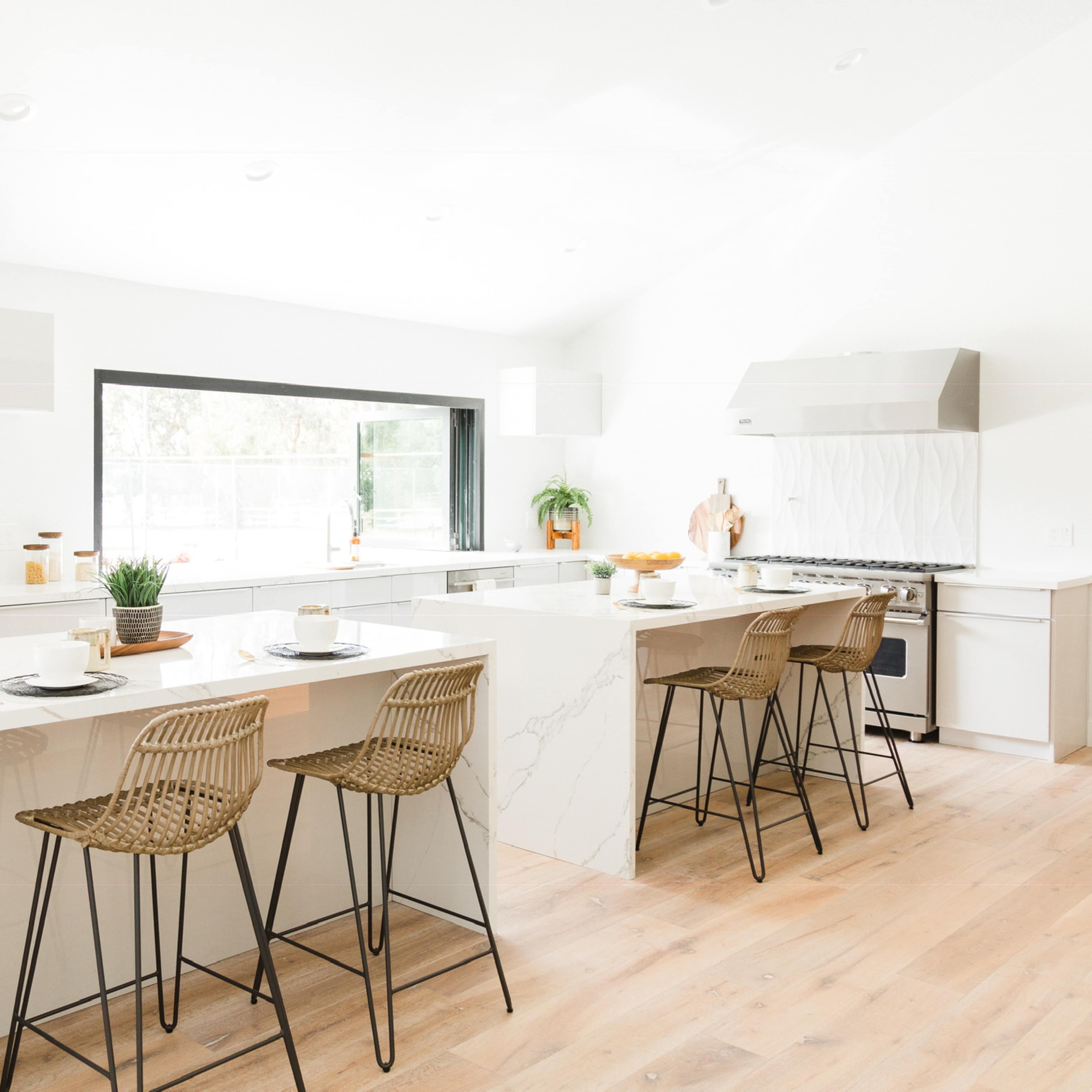 A modern kitchen features a large island with four bar stools, stainless steel appliances, and ample natural light from large windows.