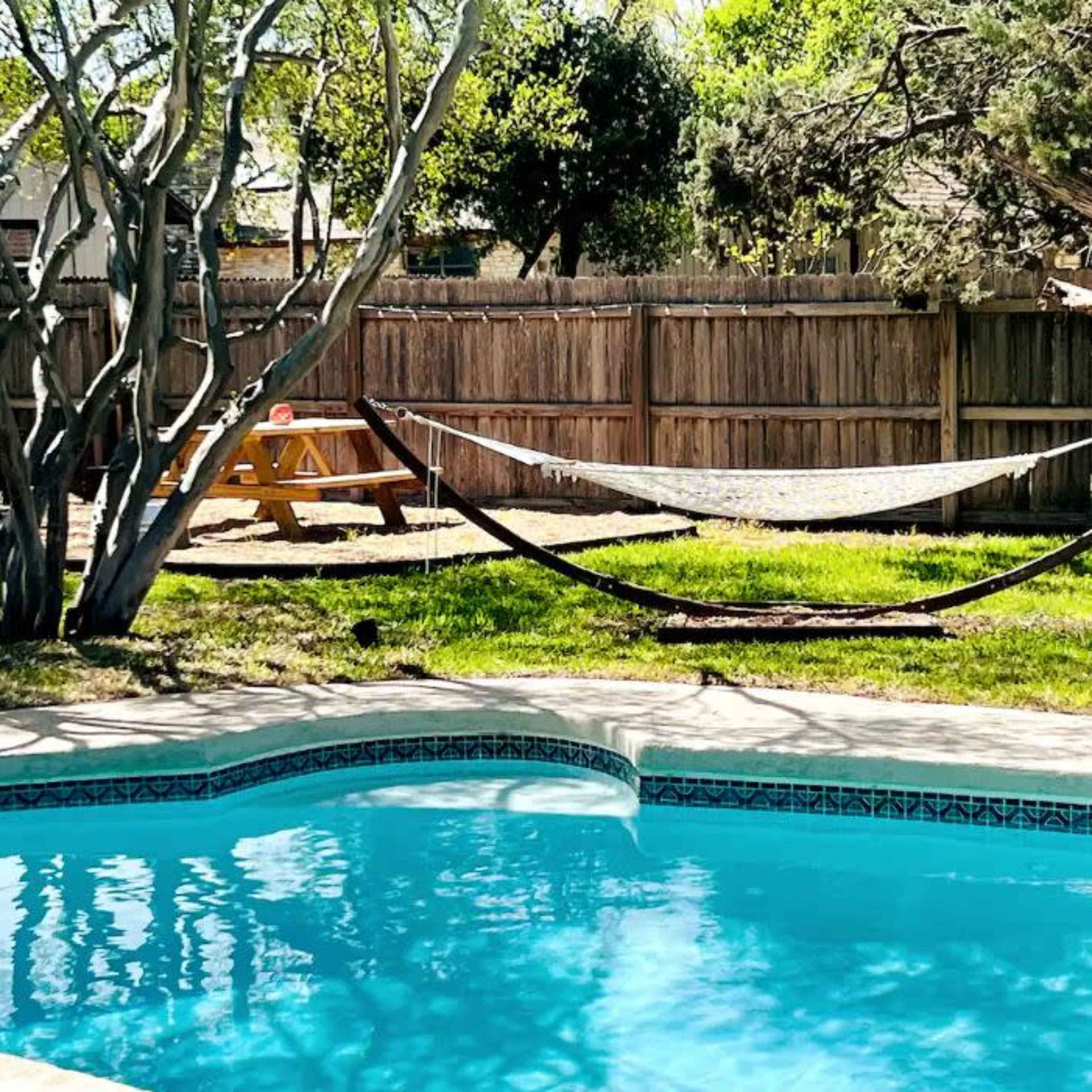 A backyard with a swimming pool, a hammock, and a picnic area surrounded by trees and a wooden fence.
