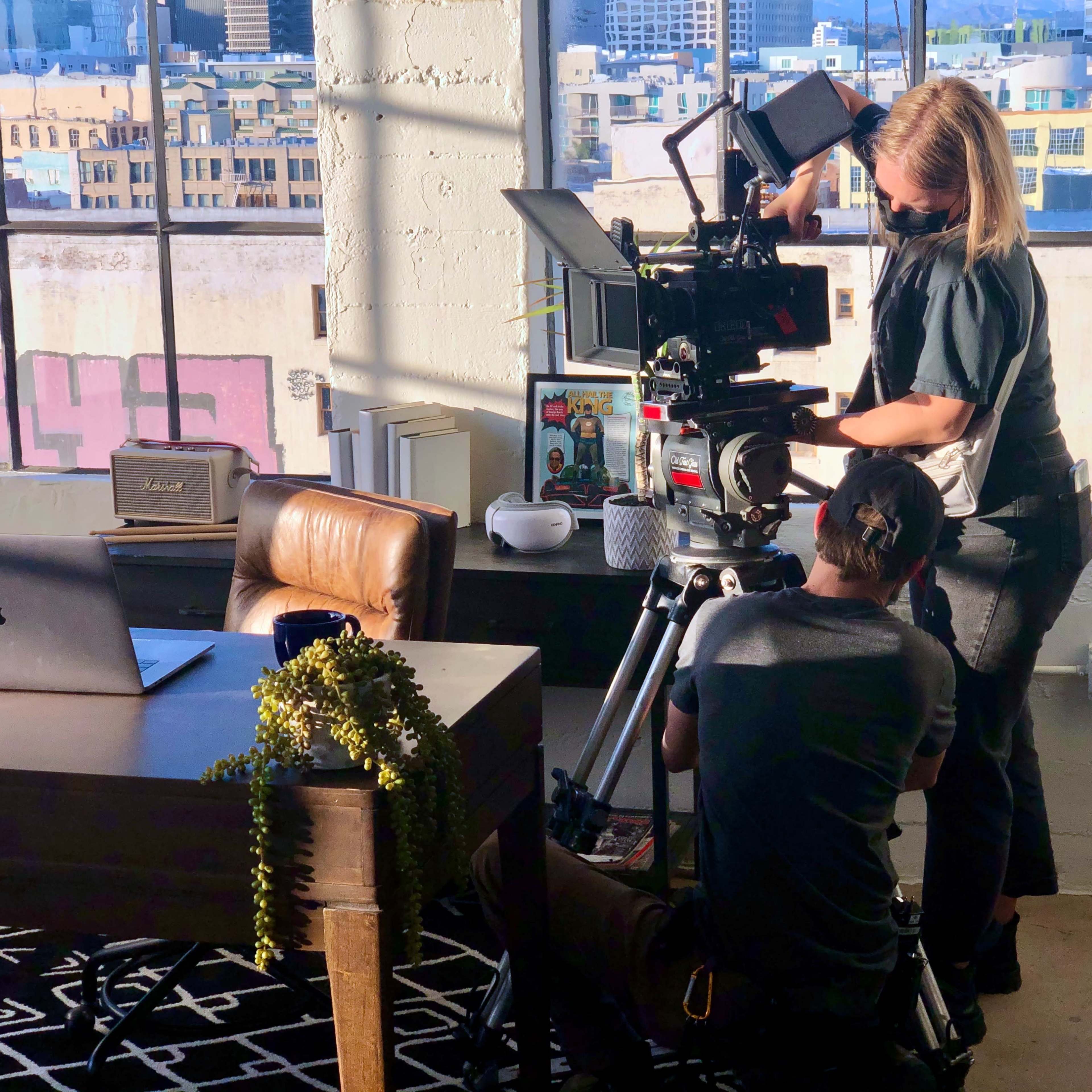 A camera crew sets up equipment in a well-lit office space with large windows overlooking a cityscape.