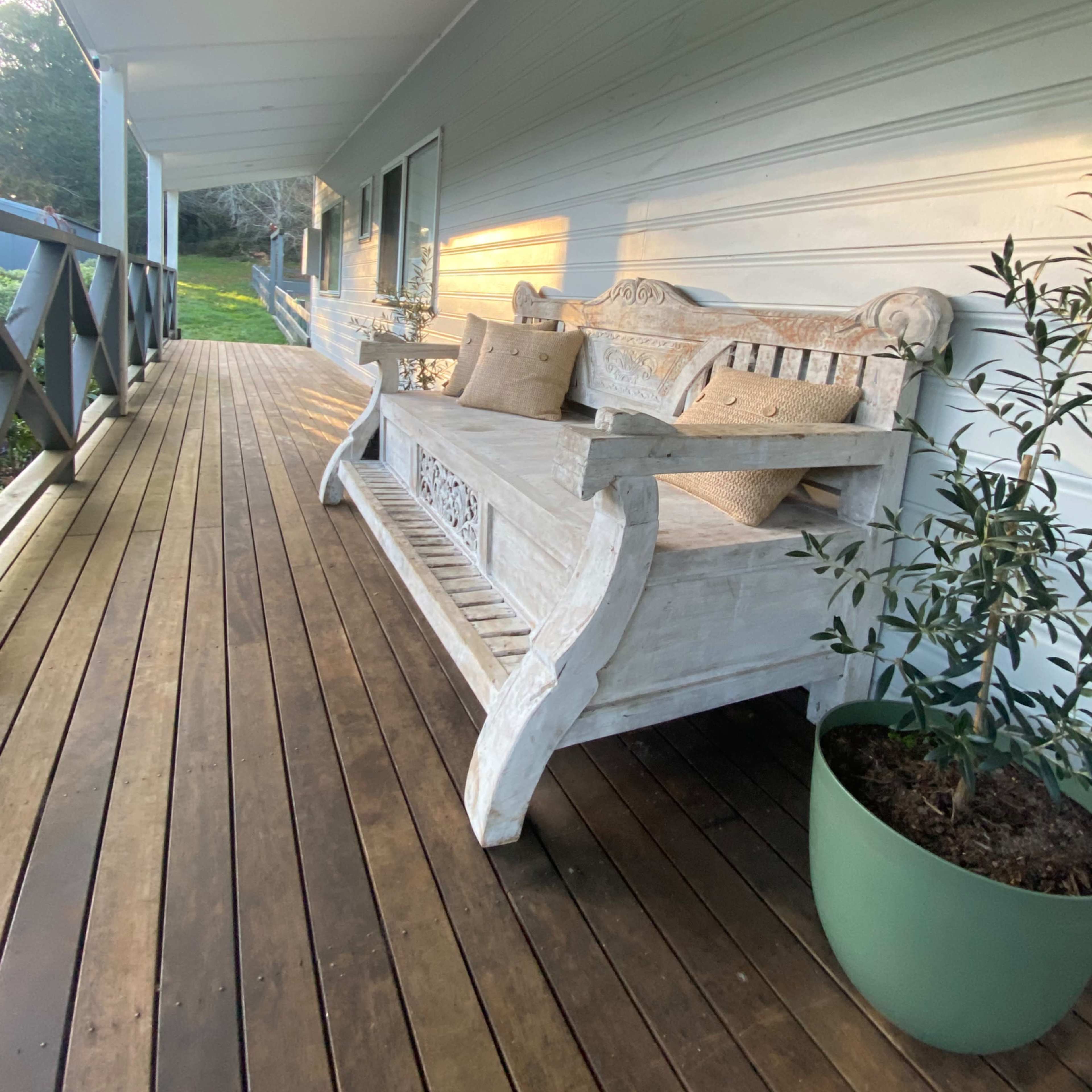 A wooden bench with decorative details is placed on a porch next to a potted plant, with sunlight illuminating the scene.