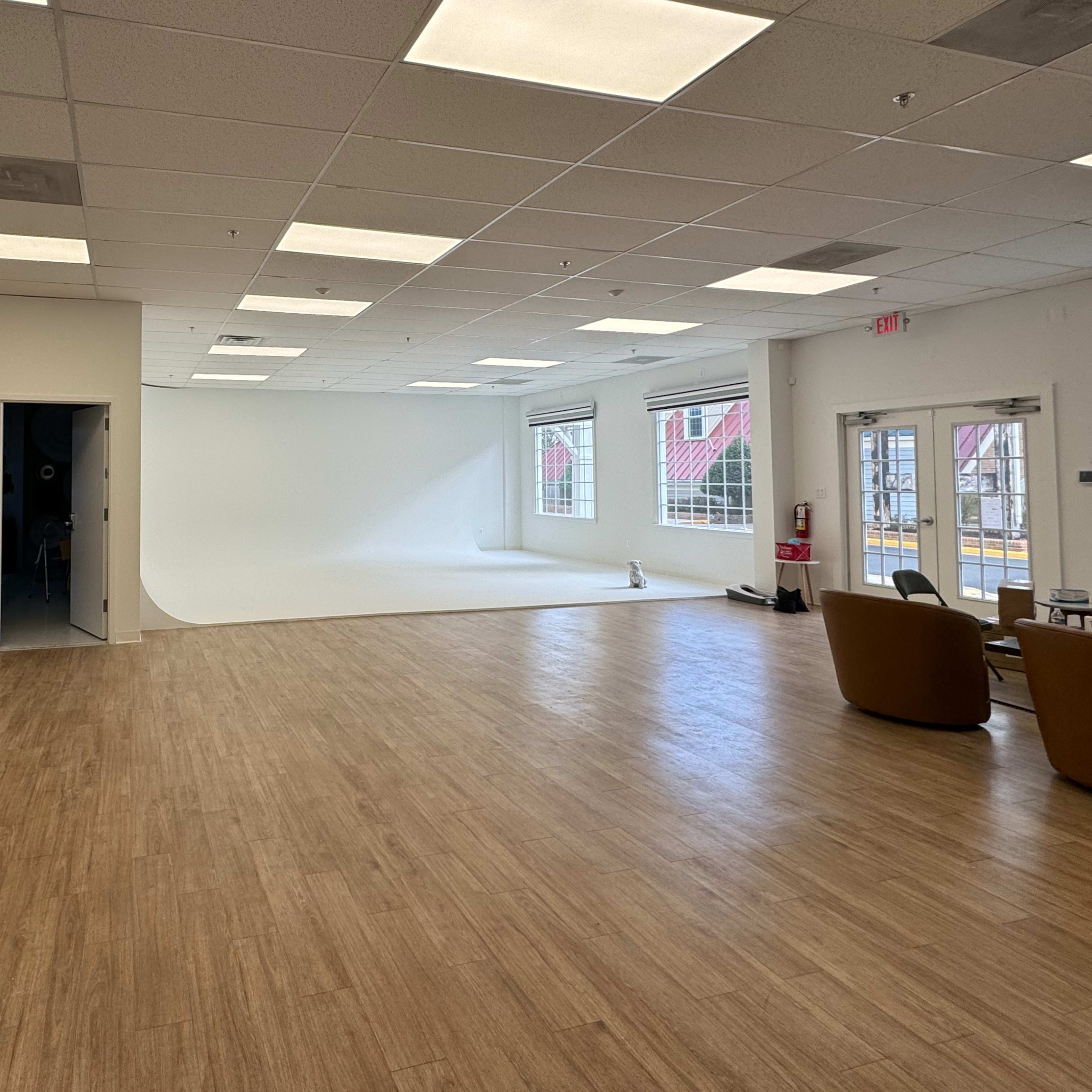 A spacious, well-lit studio with a seamless white backdrop, wooden flooring, and minimal furniture on one side.