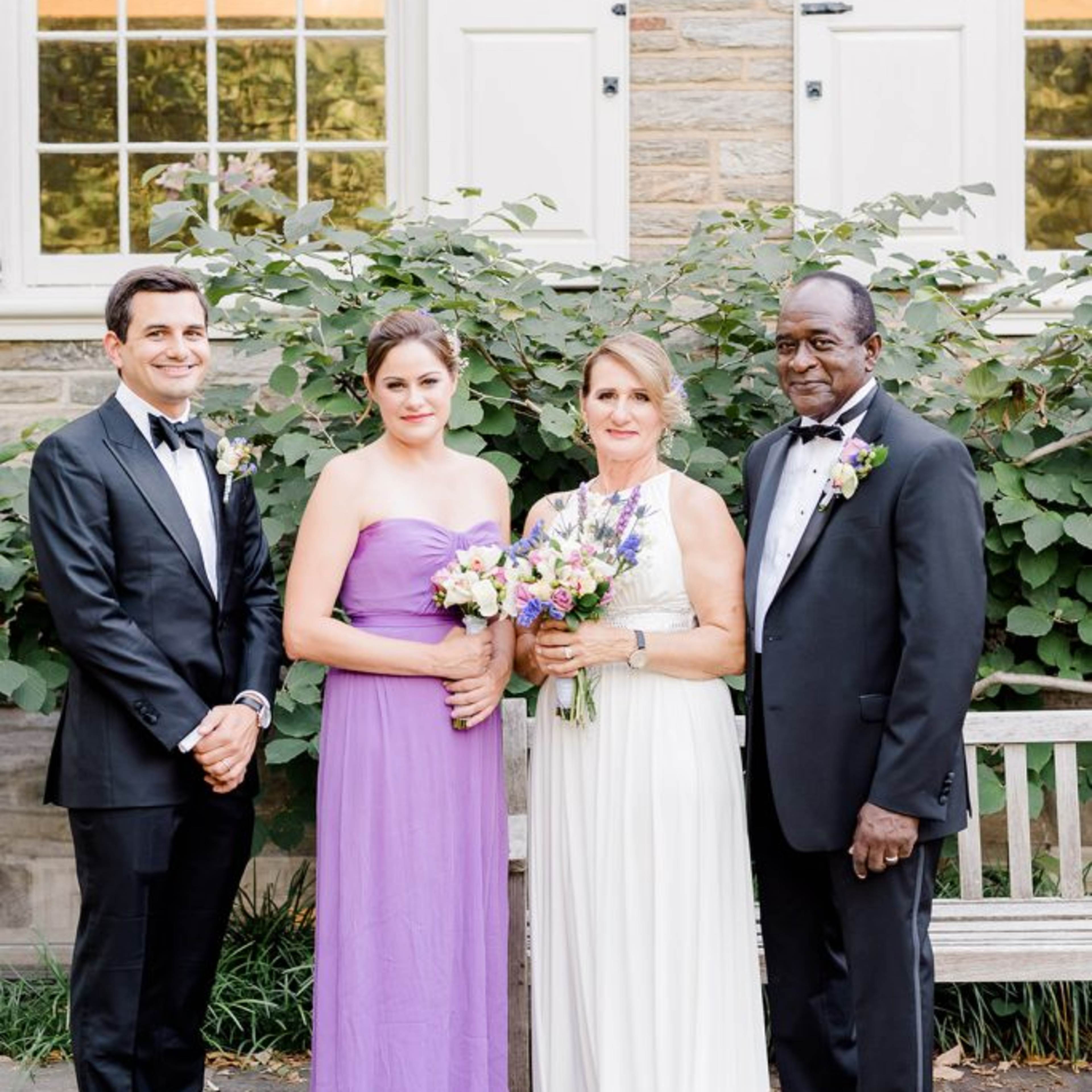 A man and a woman in formal attire stand beside two women in dresses, all posing outdoors in front of a stone building with large windows and greenery.