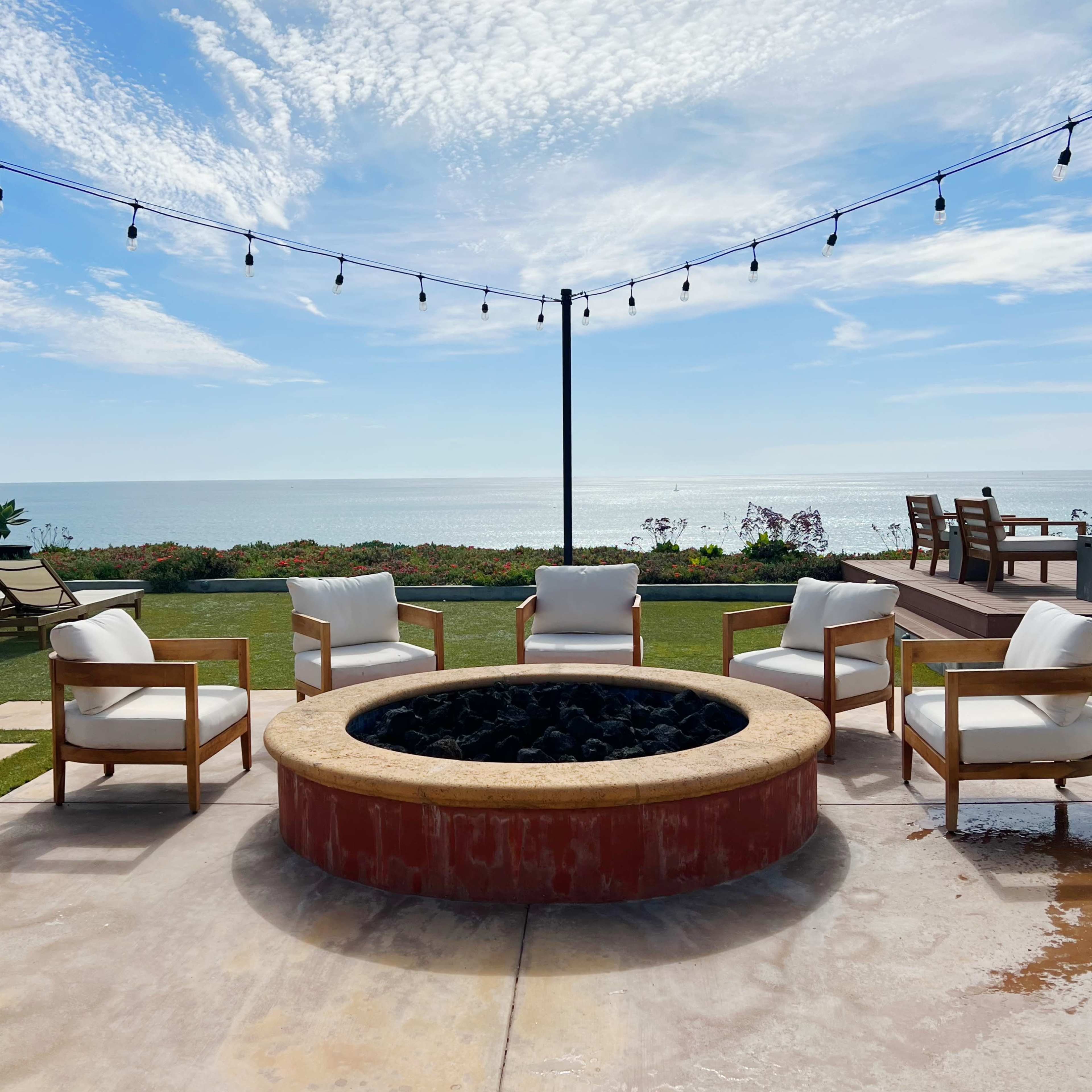 A circular fire pit surrounded by four white chairs is positioned near a coastline with a view of the ocean and a cloudy sky overhead.