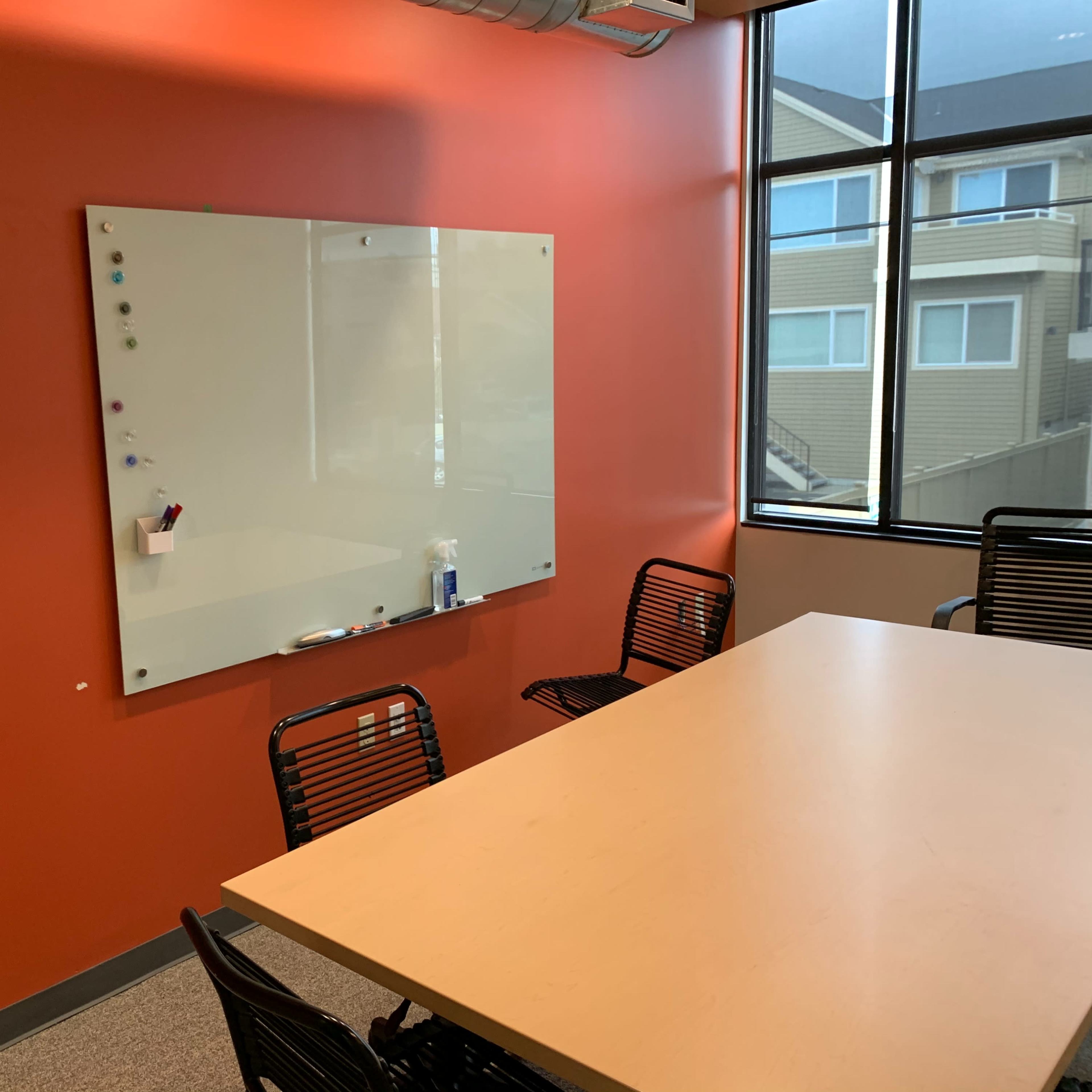 A conference room with a large whiteboard on an orange wall and a light-colored table surrounded by black chairs.