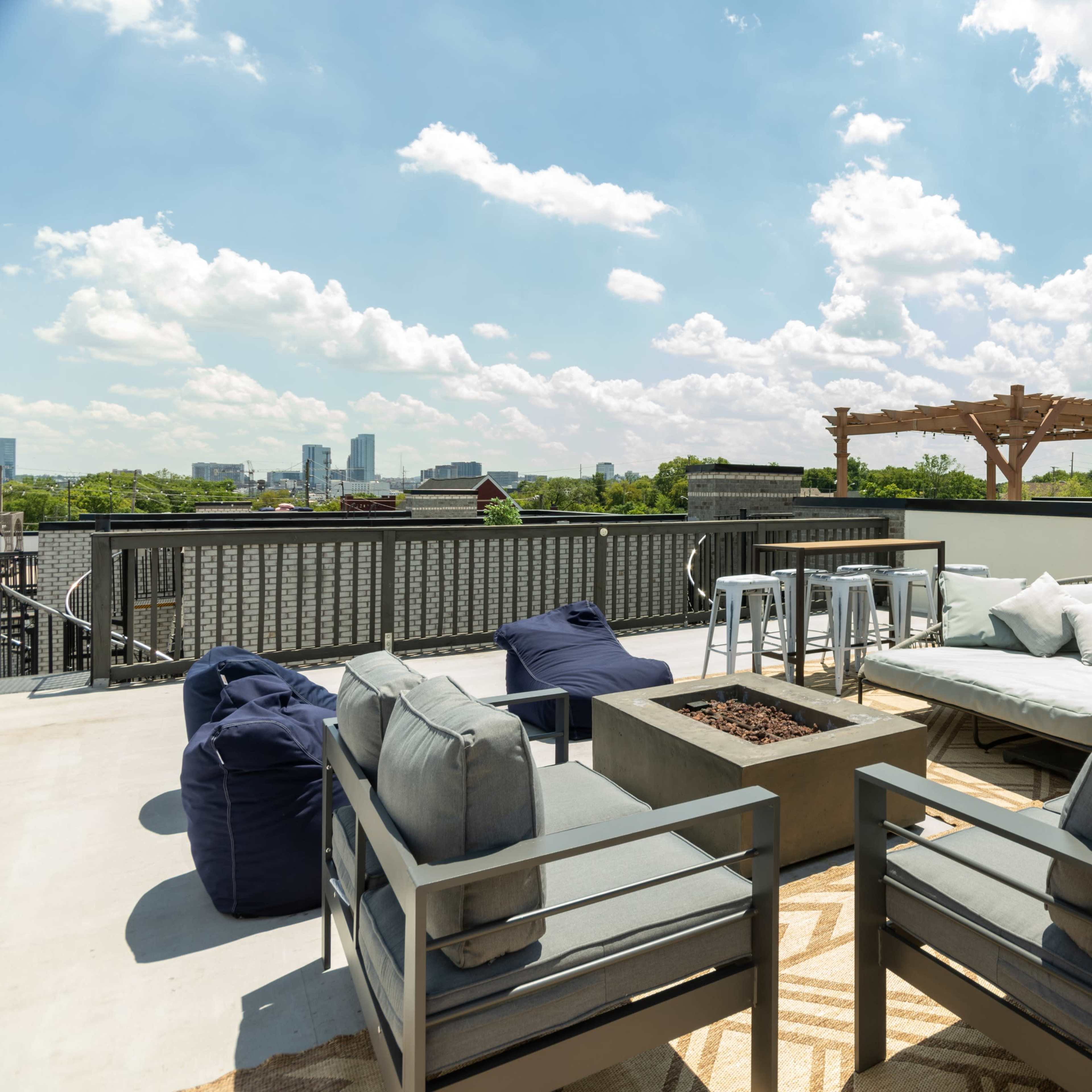 The rooftop features seating areas with grey couches and bean bags, a fire pit, and city views under a blue sky with scattered clouds.