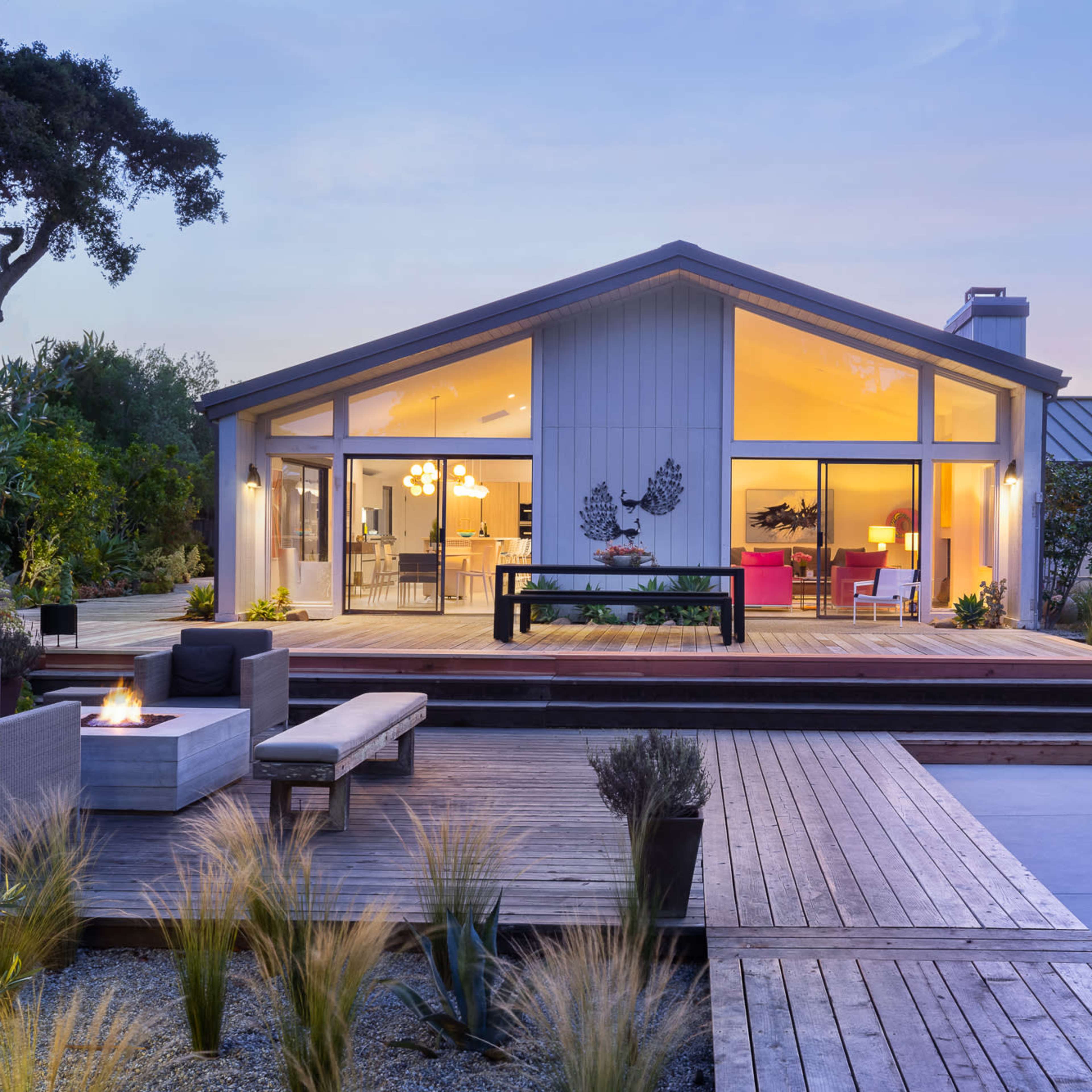 A modern, single-story house with large windows and a wooden deck features a fire pit and landscaped surroundings, set against a twilight sky.