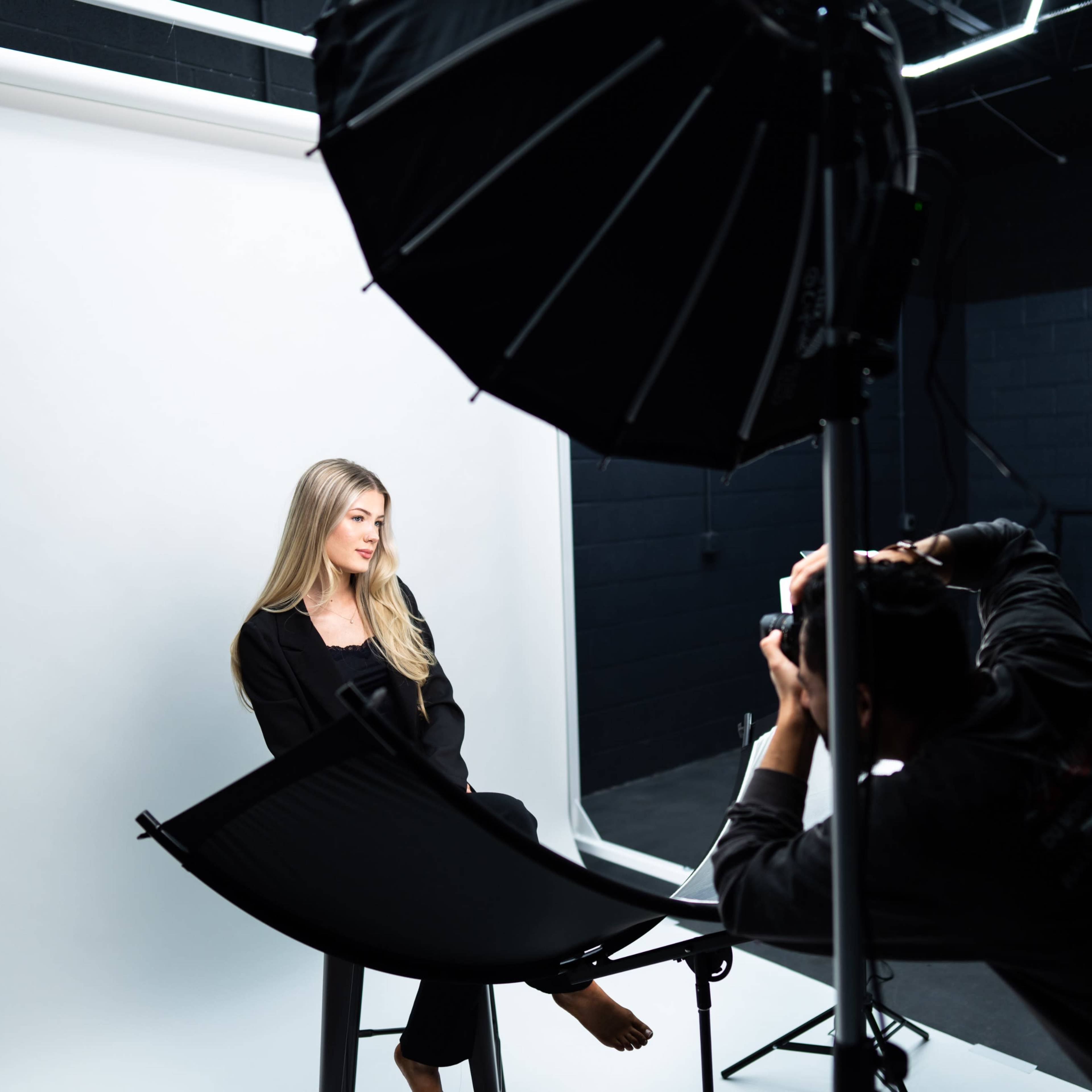 A photographer captures a model seated in a studio with a plain backdrop and professional lighting setup.