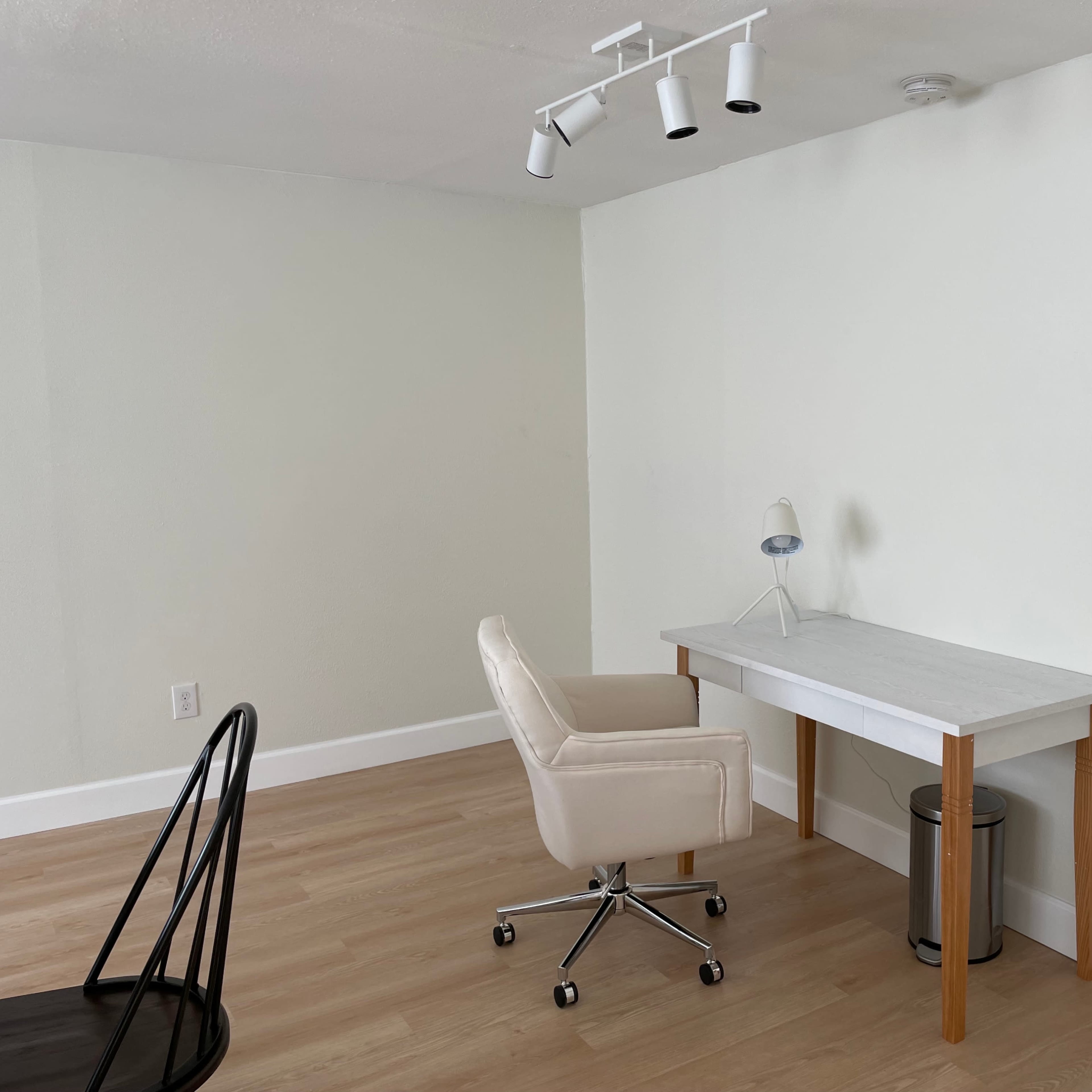 A minimalist workspace featuring a white desk with a lamp, a swivel chair, and a waste bin against a light-colored wall.