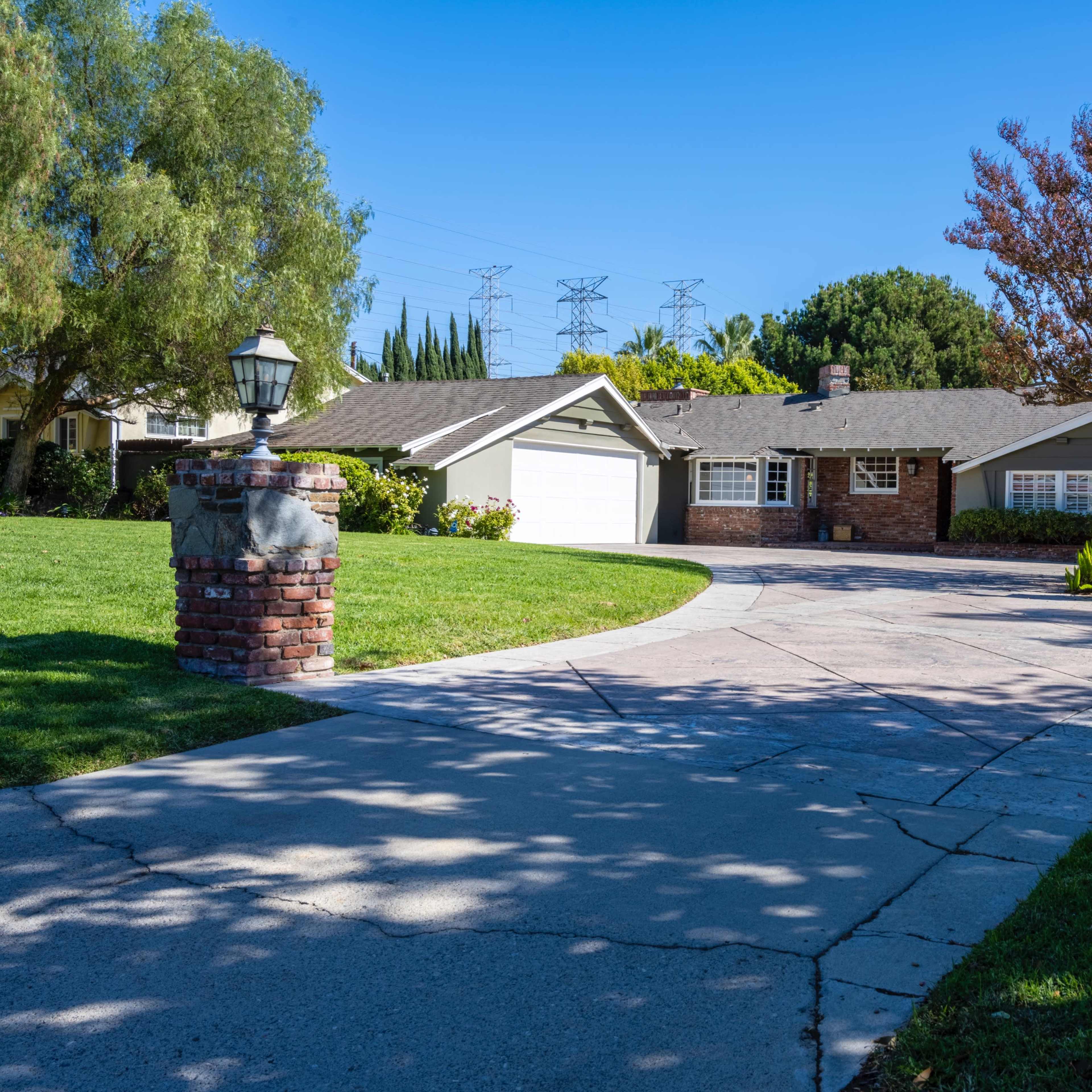 A single-story suburban house with a front lawn, a wide driveway, and a tree with red leaves.