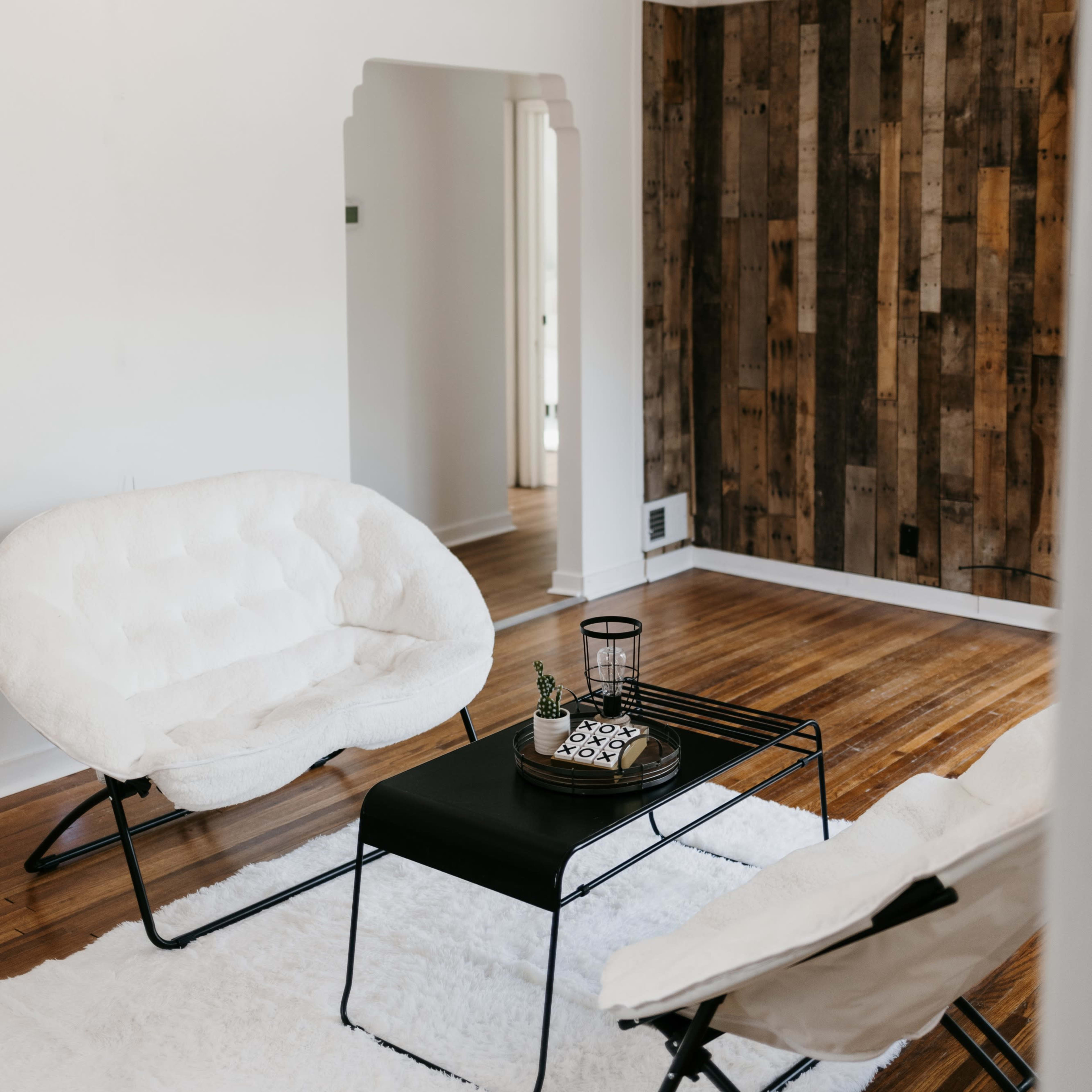 A living room with two white chairs, a black table, and wood-paneled walls, illuminated by string lights overhead.