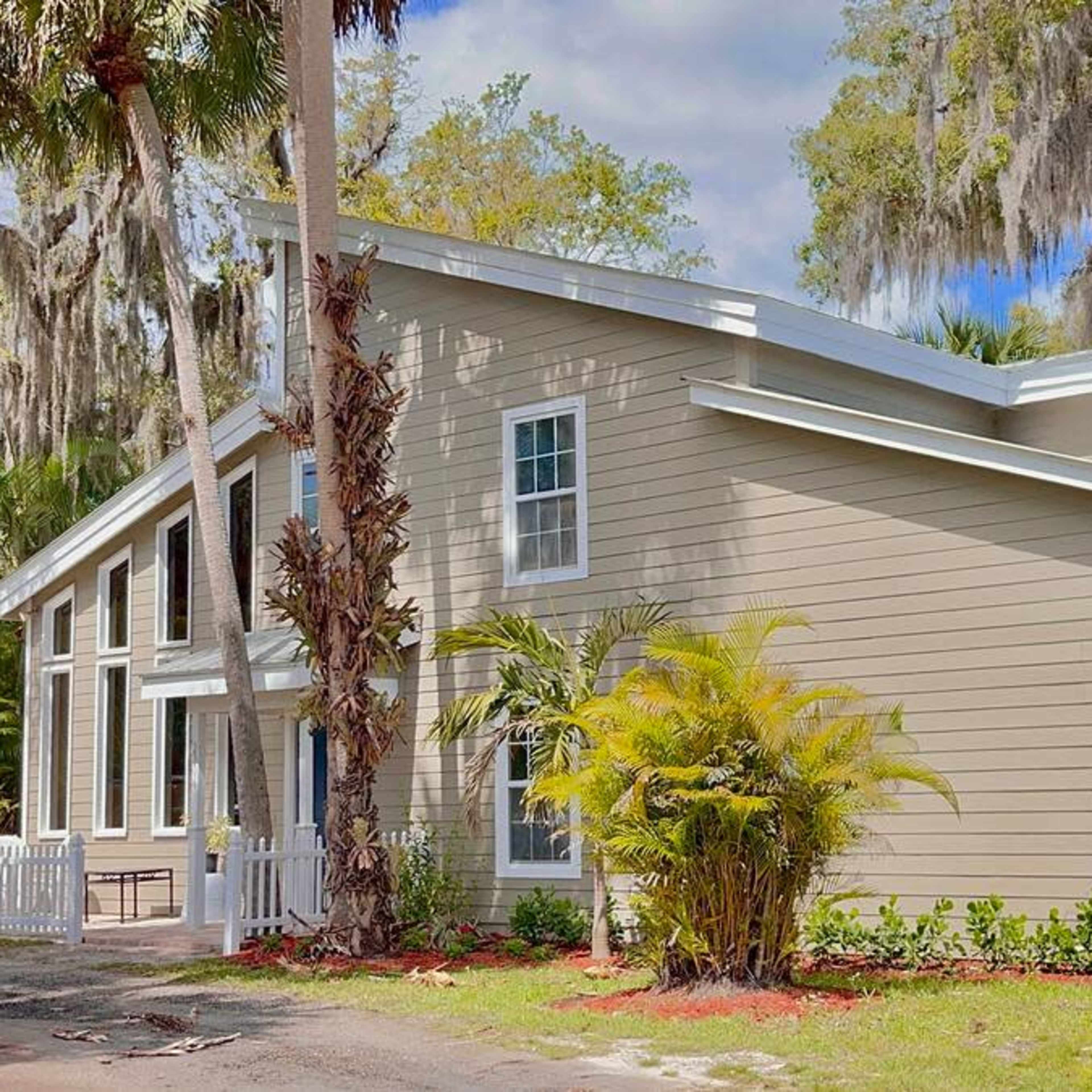 A light-colored house with large windows and palm trees, located in a landscaped area with a gravel path.
