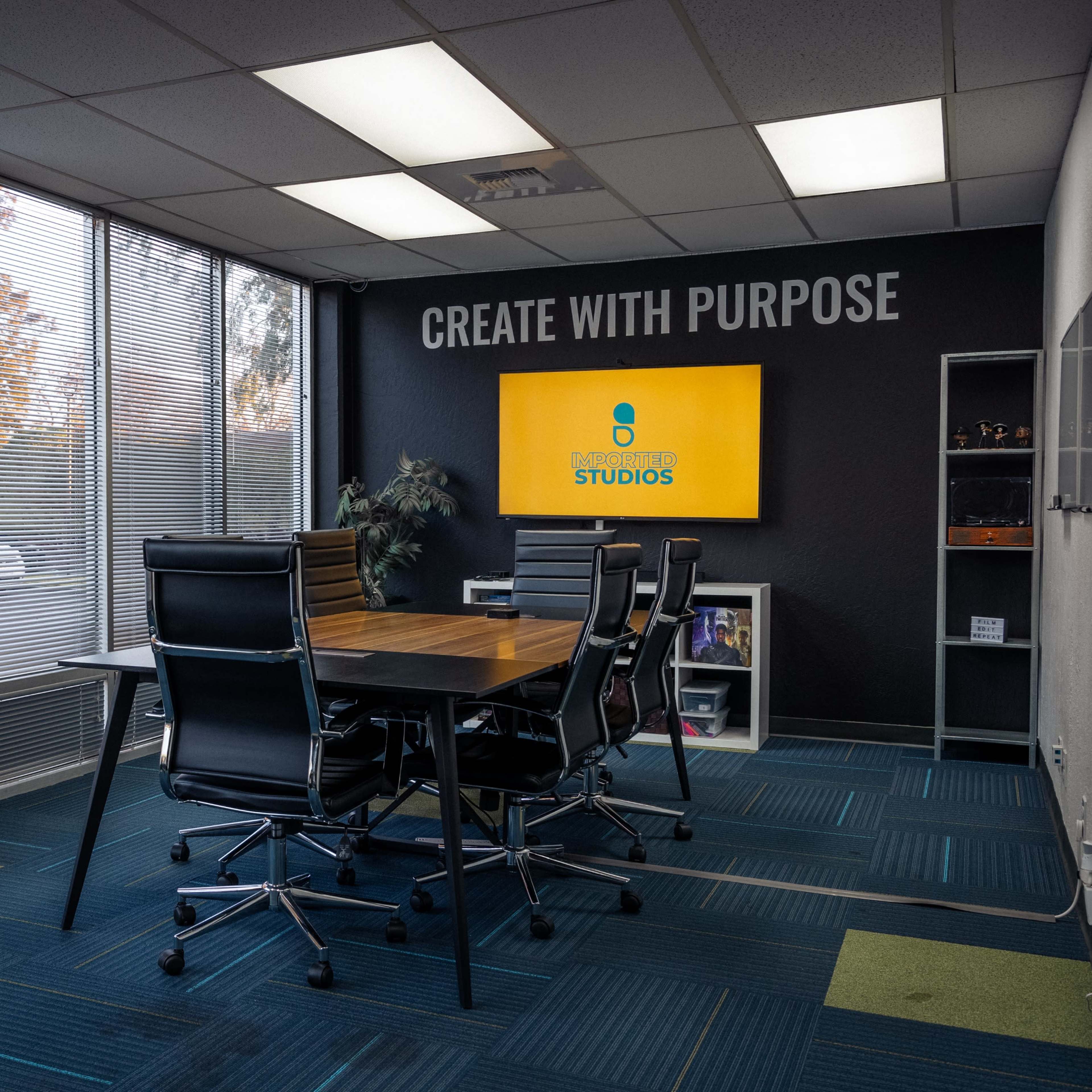 A conference room features a table surrounded by black chairs, large windows letting in daylight, and a wall with the phrase "CREATE WITH PURPOSE" and a screen displaying the name "STUDIOS."