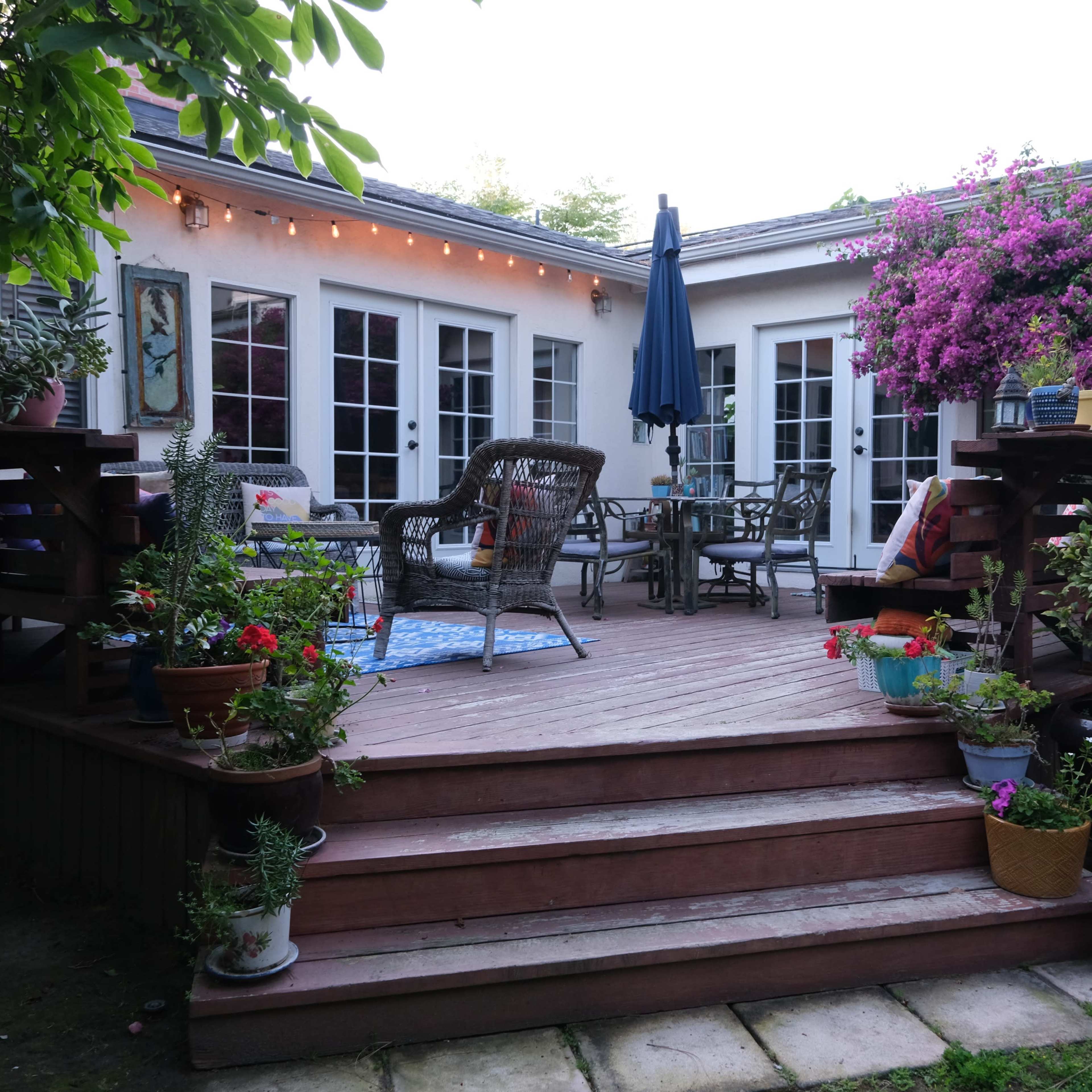 A wooden deck surrounded by potted flowers and plants, leading to a seating area with a table and chairs, illuminated by string lights.