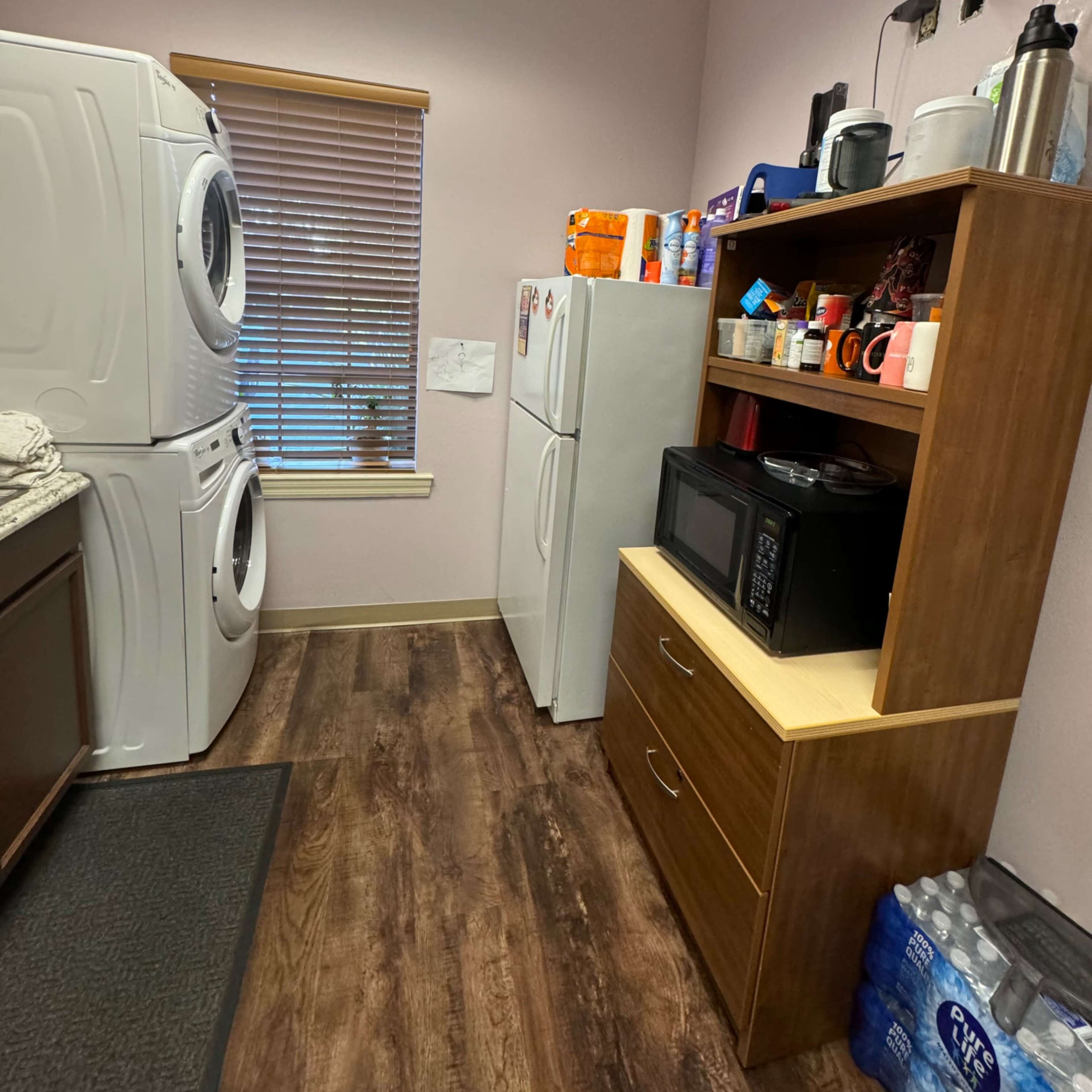 A laundry room featuring a stacked washer and dryer on the left, a refrigerator next to them, and a wooden shelf with a microwave and various items on the right.