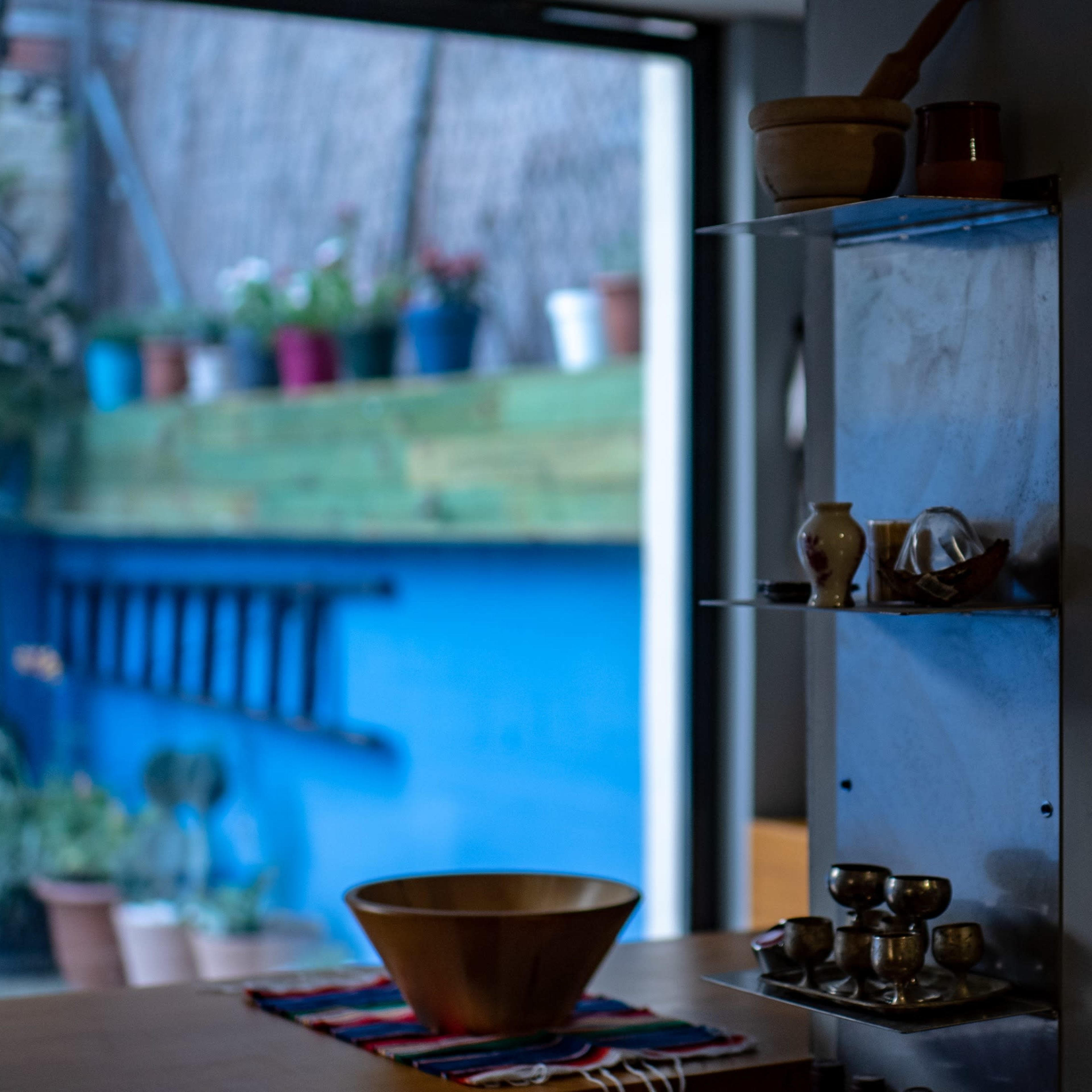 A modern kitchen interior with a wooden bowl on a table and a view of a colorful outdoor area filled with potted plants.