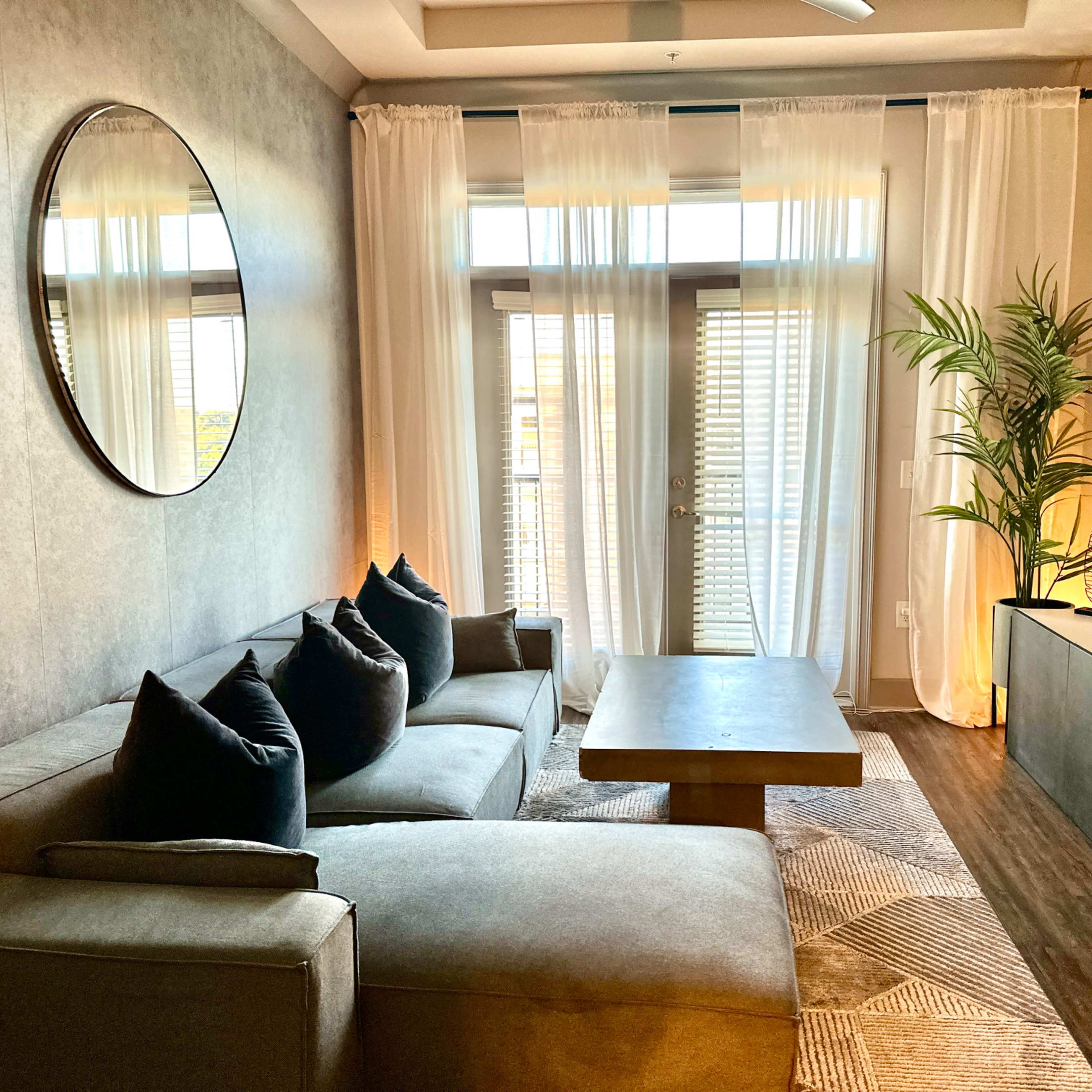 A modern living room features a sectional sofa, a round mirror, large windows with sheer curtains, a flat-screen TV on a cabinet, and a potted plant.
