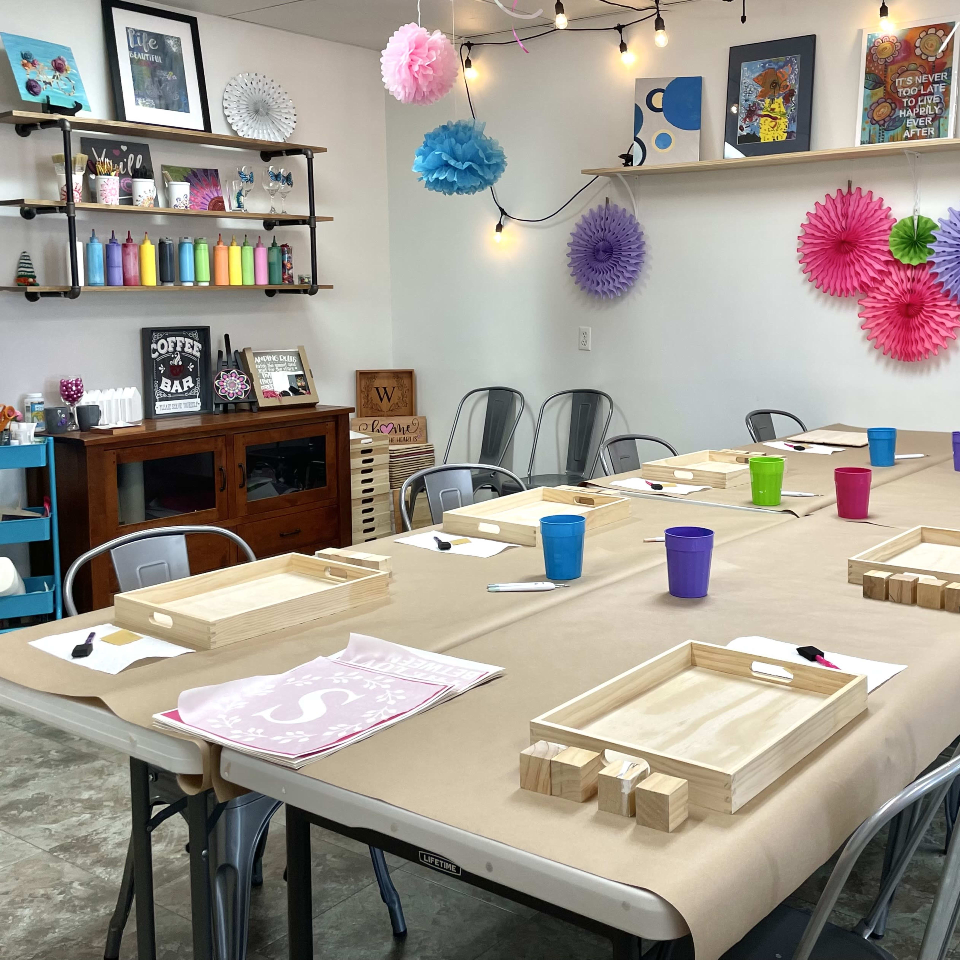 A large, rectangular table is set for an event, covered with brown paper, and surrounded by metal chairs, with colorful cups and wooden trays arranged neatly at each place, and decorations hanging on the walls.