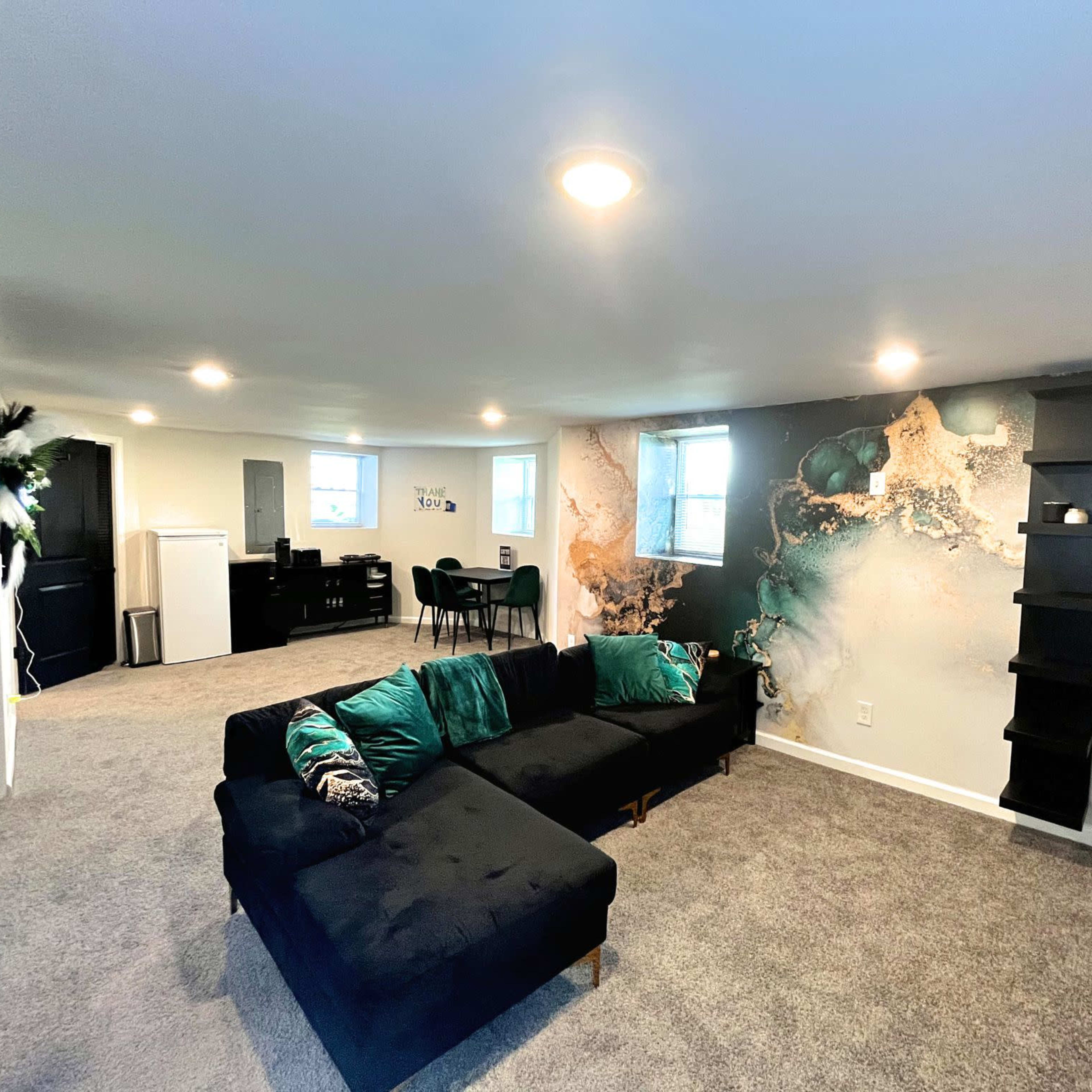 A spacious living area featuring a black sectional sofa, a decorative wall with a multi-colored design, and a dining area with a table and chairs in the background.