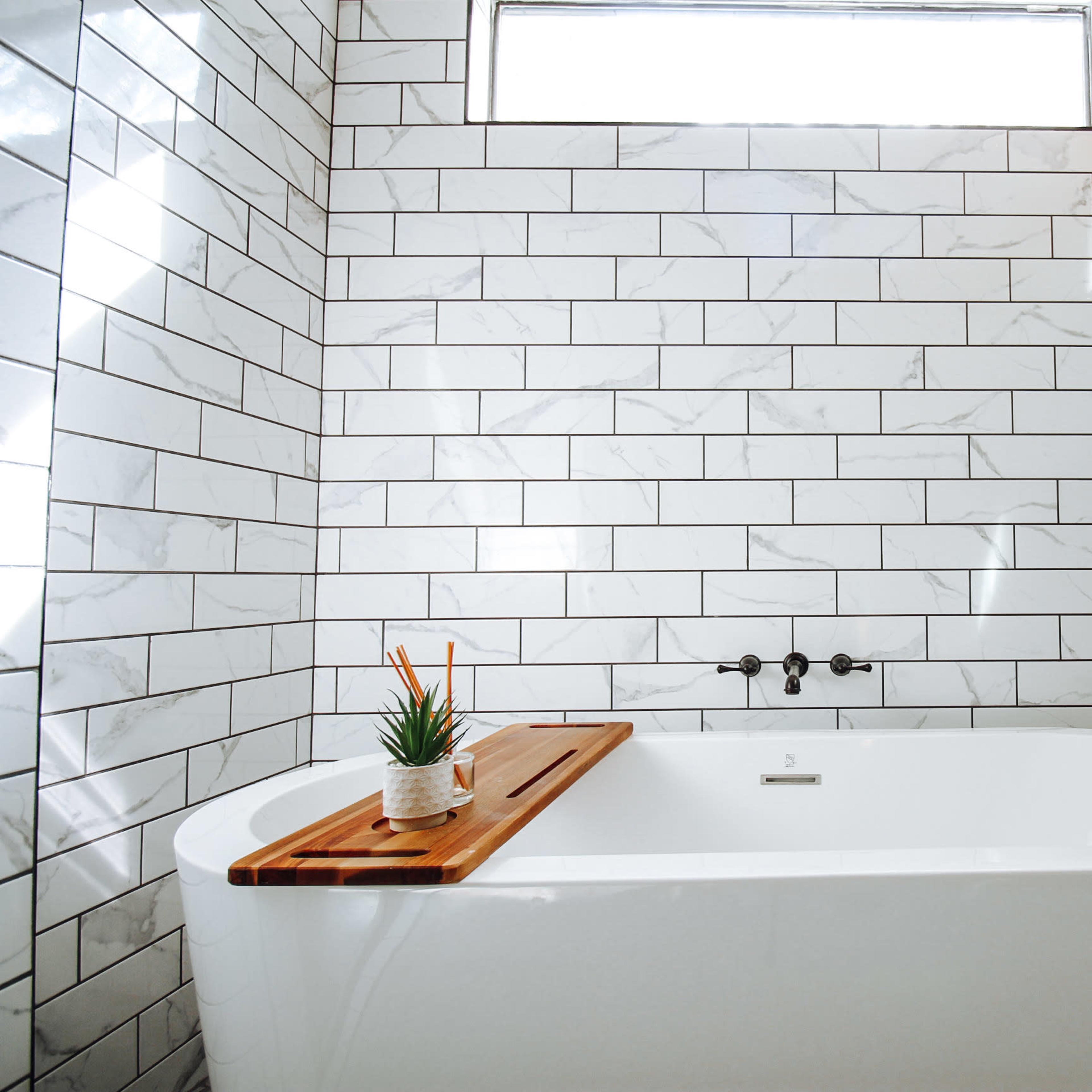 A modern bathroom with a freestanding white bathtub, a wooden bath tray, and tiled walls featuring a mix of white and marble patterns.
