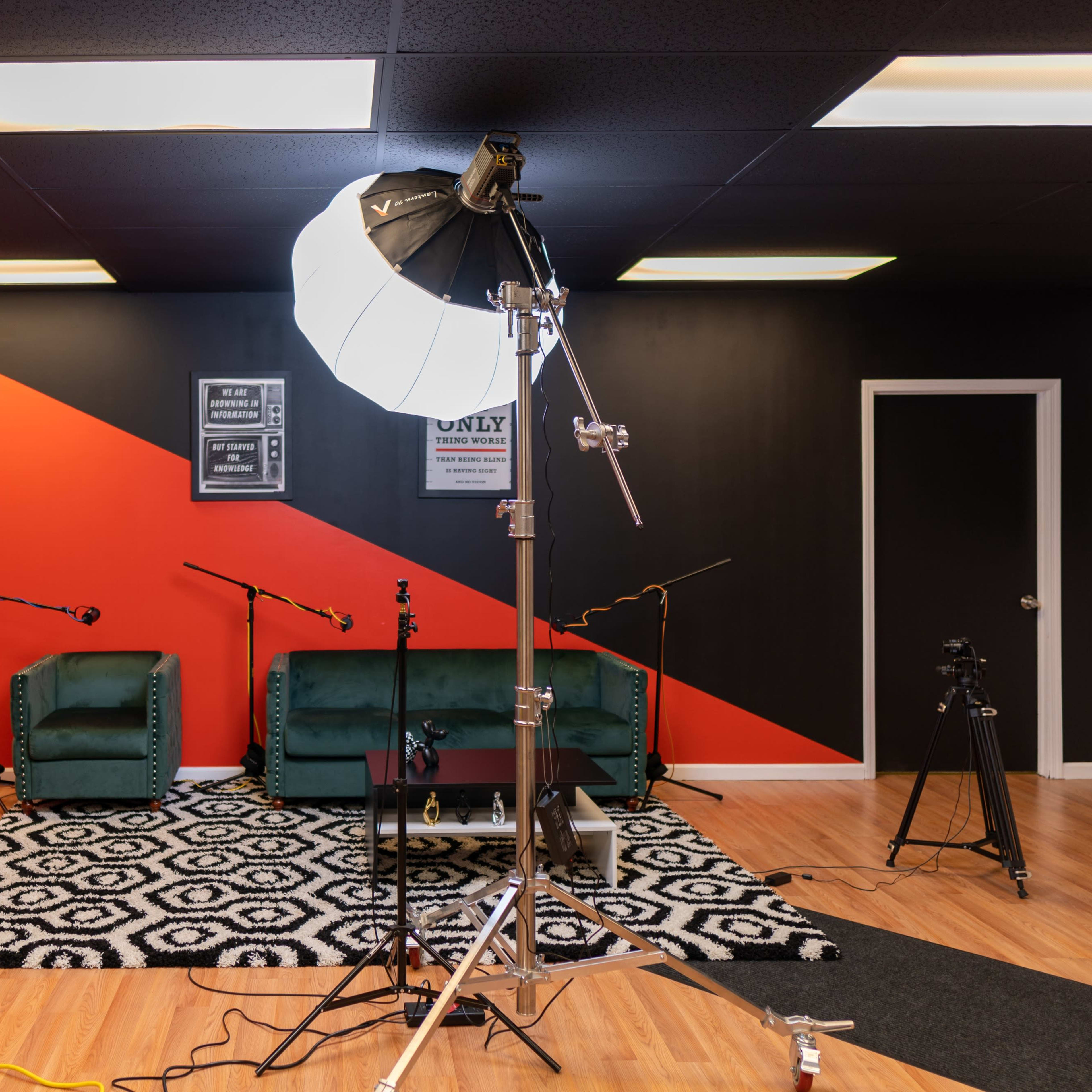A modern studio setup featuring green velvet chairs, a patterned rug, lighting equipment, and microphones against a backdrop of black and red walls.