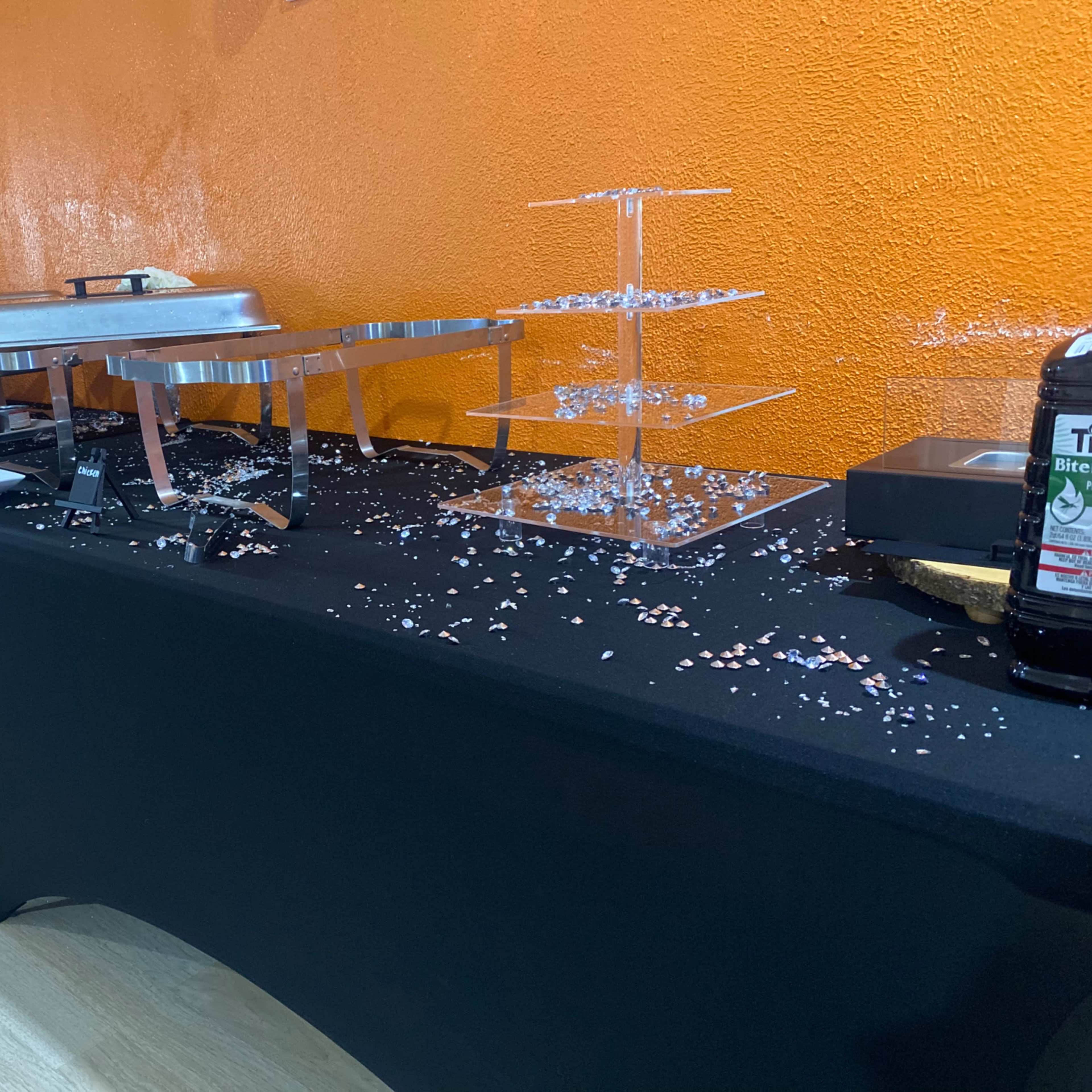 A buffet setup features chafing dishes and clear display stands on black tablecloths, with decorative confetti scattered across the surface.