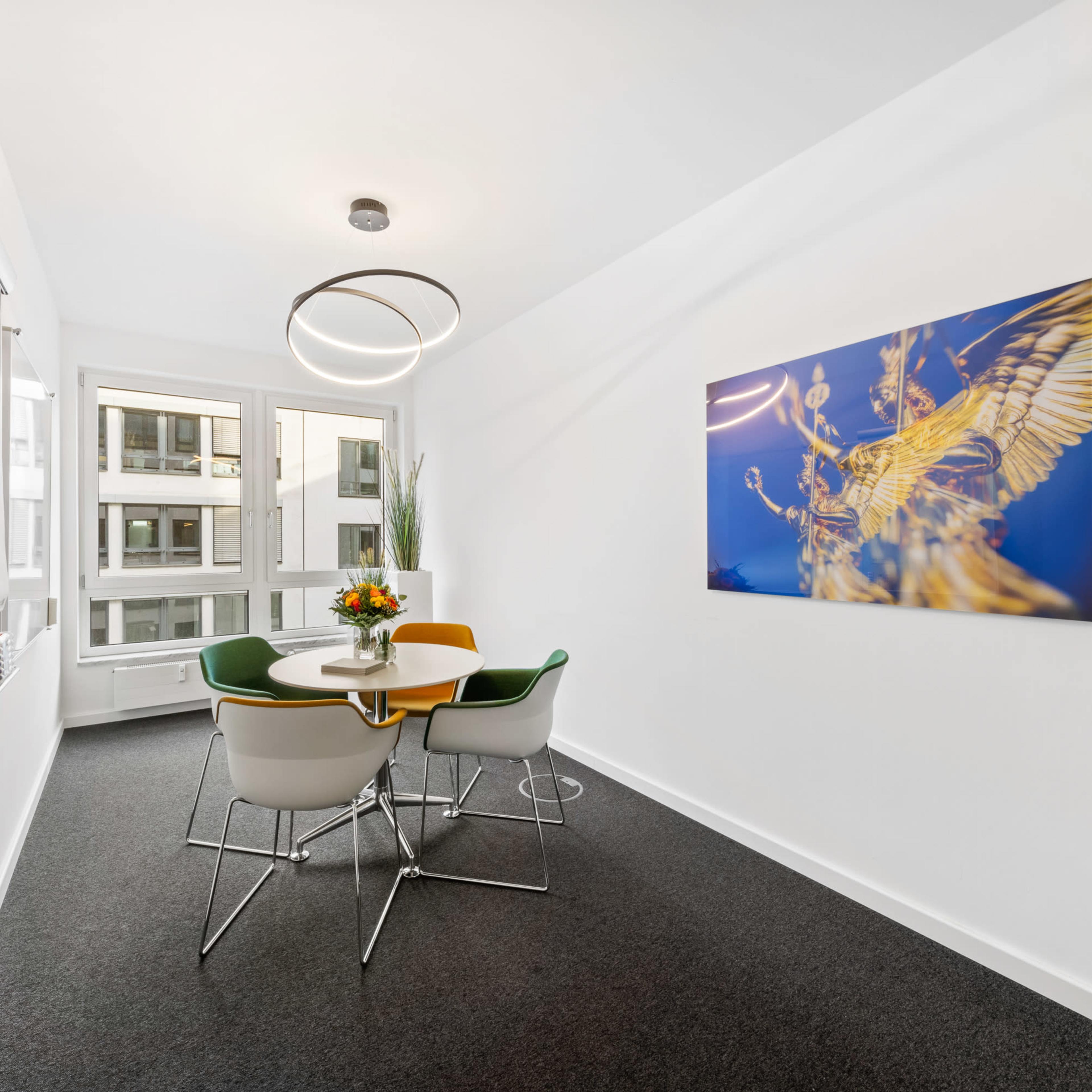A small, modern meeting room features a round table with four chairs and a large decorative wall art piece.