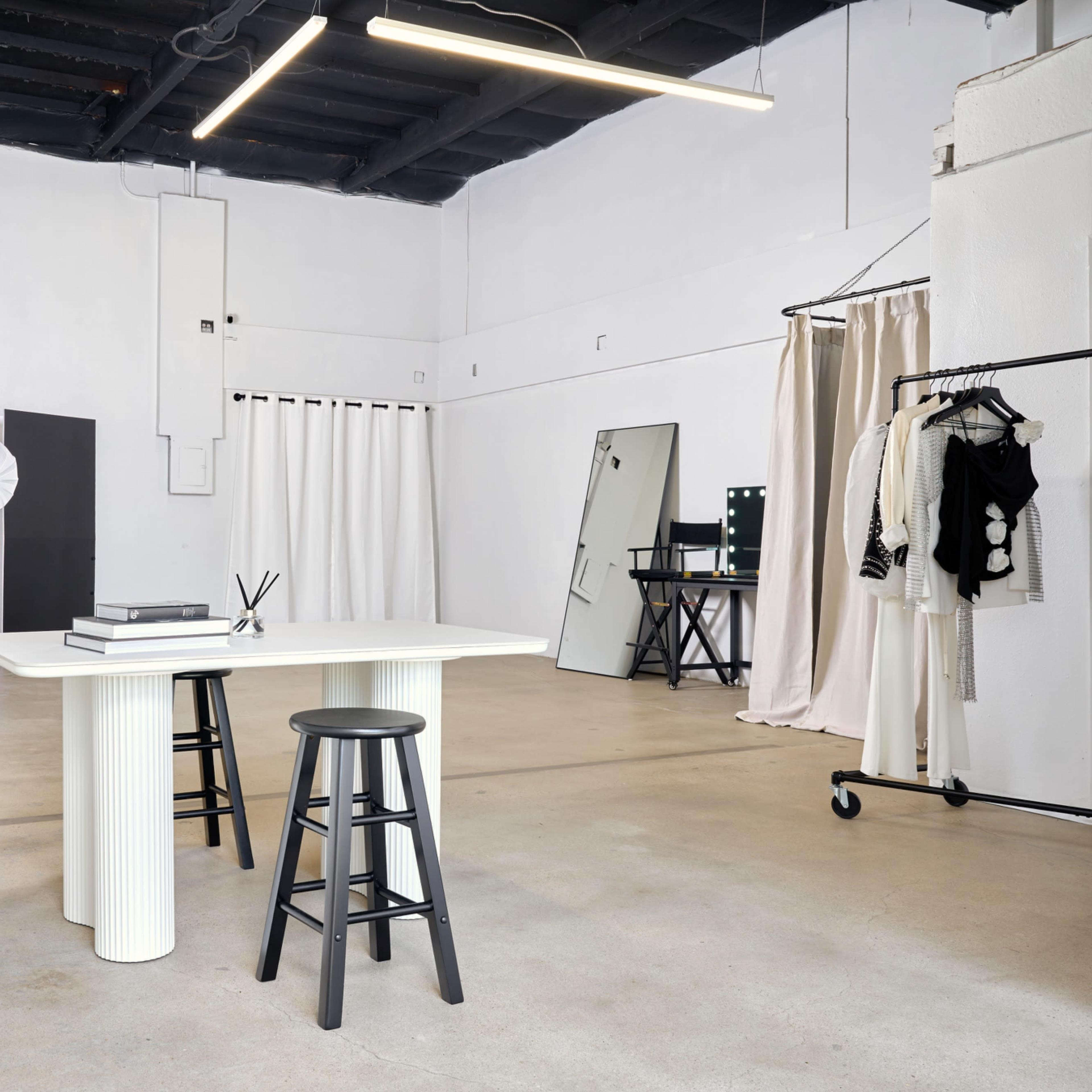 A minimalist studio space with a white wall, two black stools, a table, and various photography equipment set against light-colored flooring.