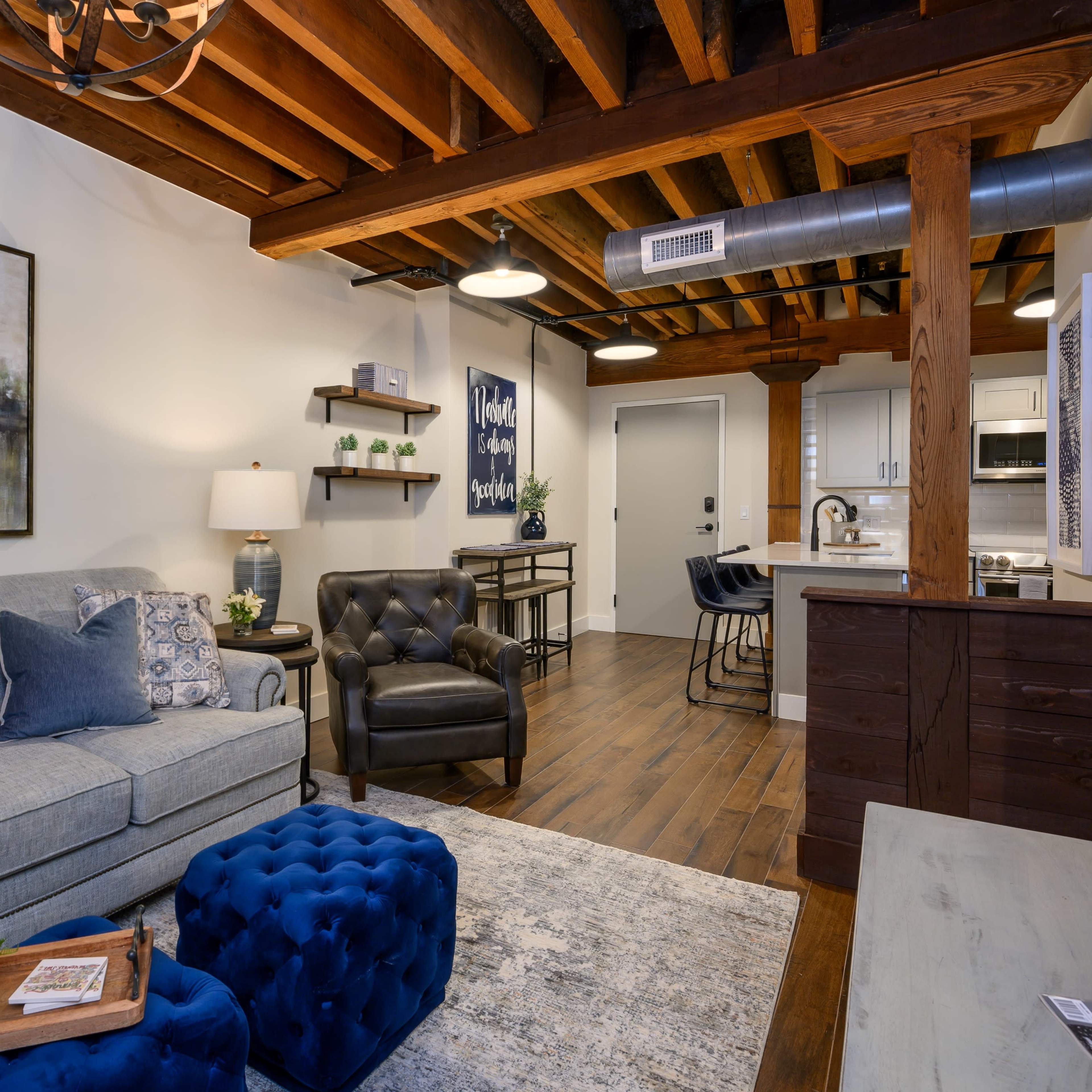 A cozy living room with a gray sofa, a blue ottoman, and exposed wooden beams in the ceiling, leading into an adjacent kitchen area.