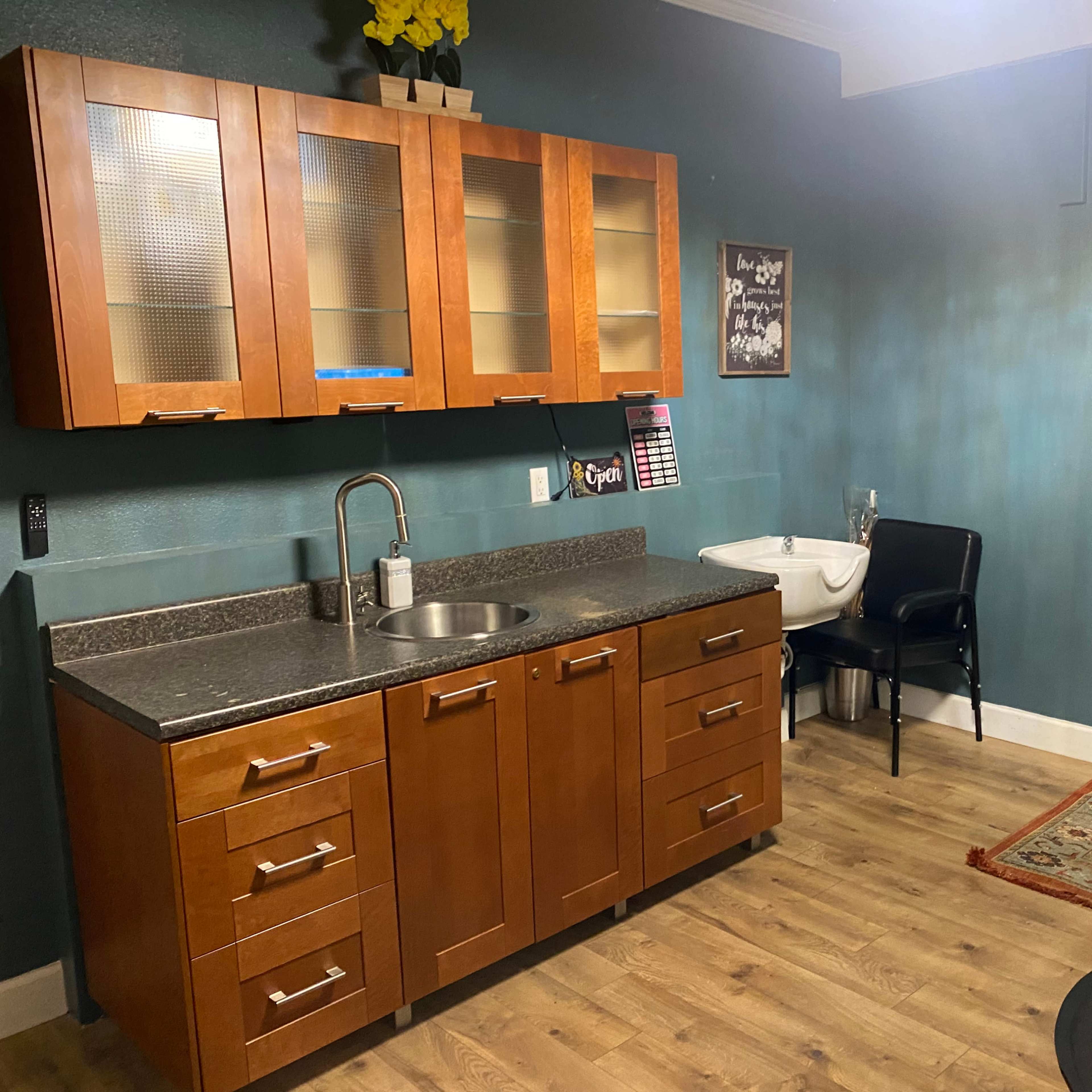 A small room features a wooden cabinetry with a sink, a black chair, and a decorative rug on a wood floor.