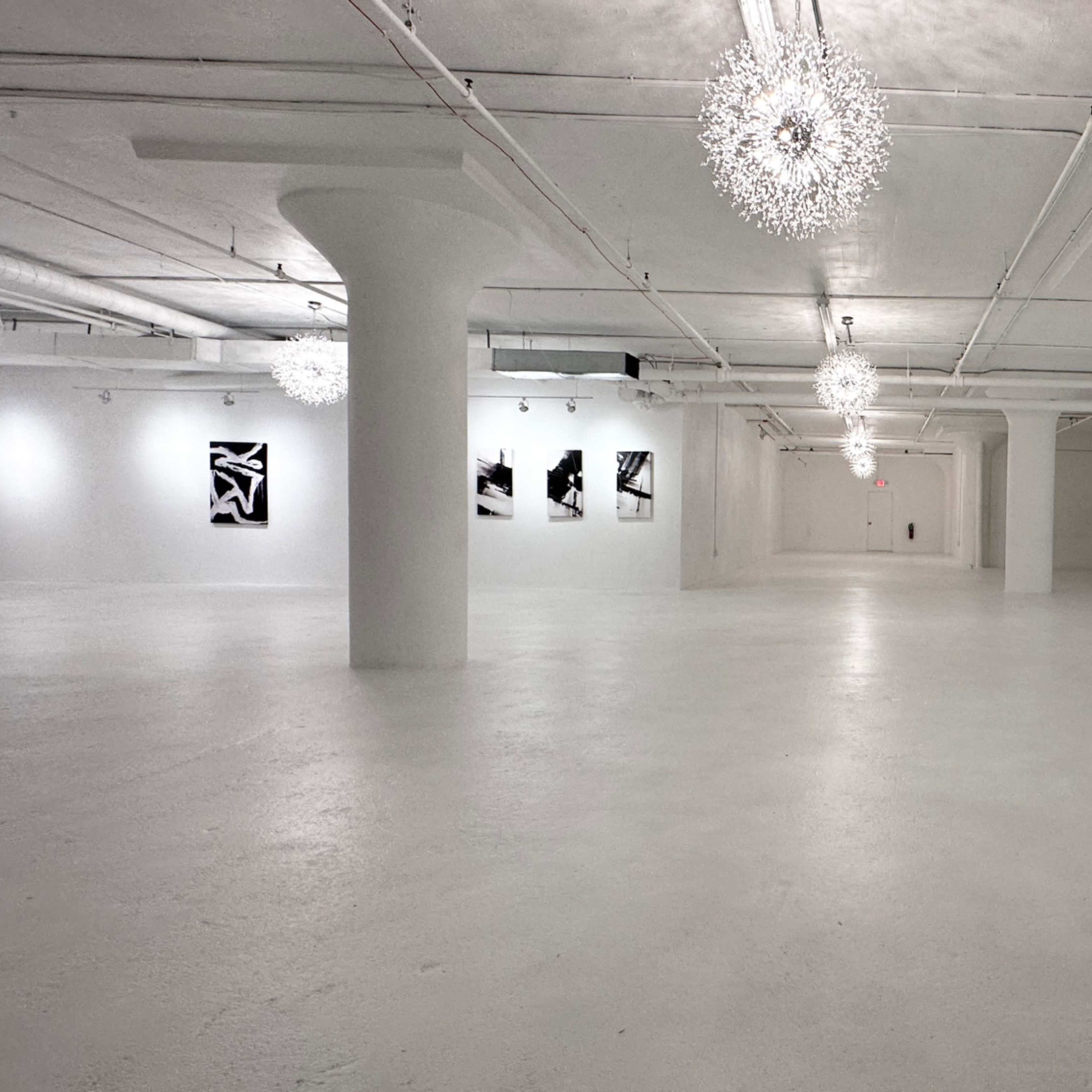 A spacious, white-walled art gallery with several black-and-white artworks displayed on the walls and decorative light fixtures hanging from the ceiling.