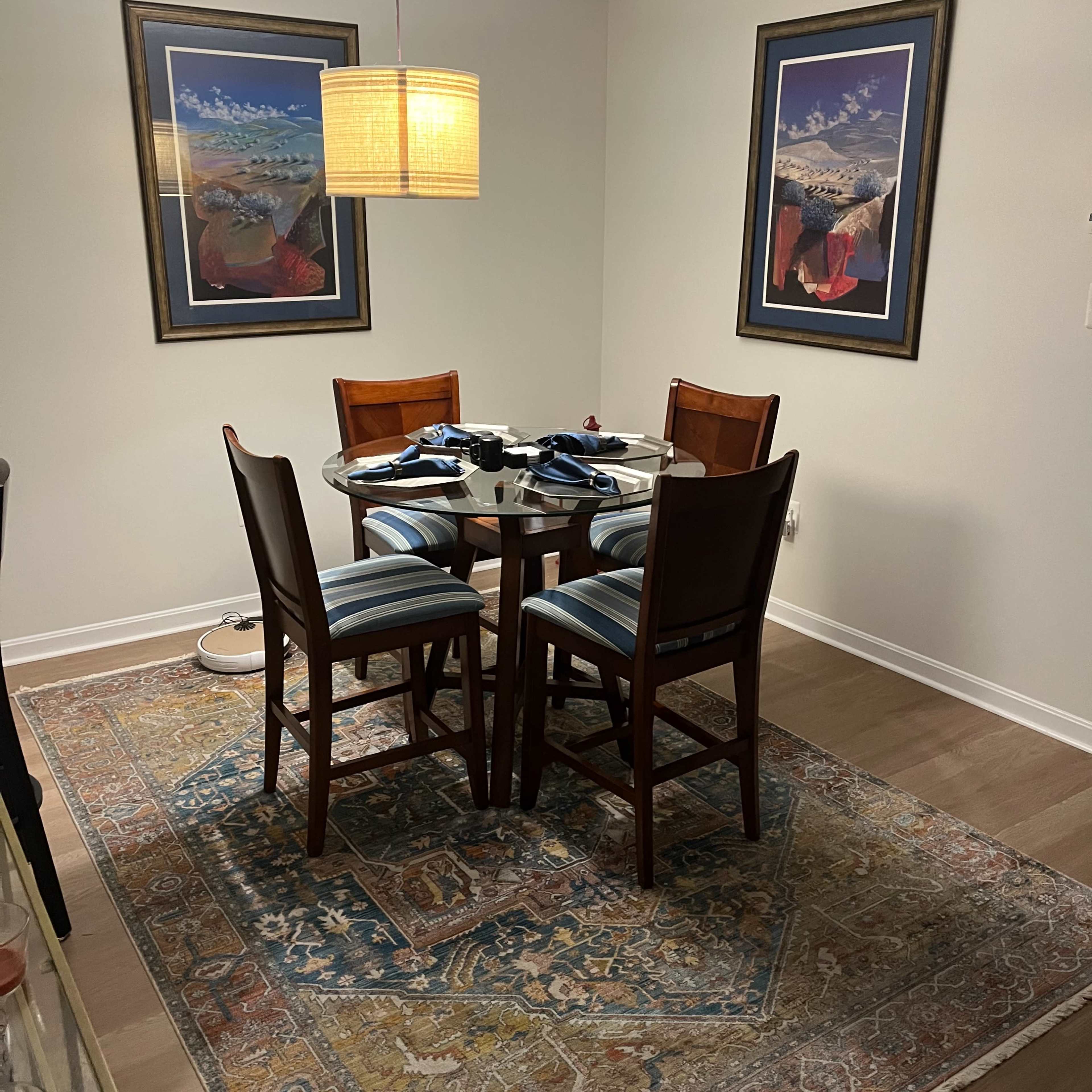 A dining area with a round glass table surrounded by four wooden chairs, set on a patterned rug, and illuminated by a pendant light.