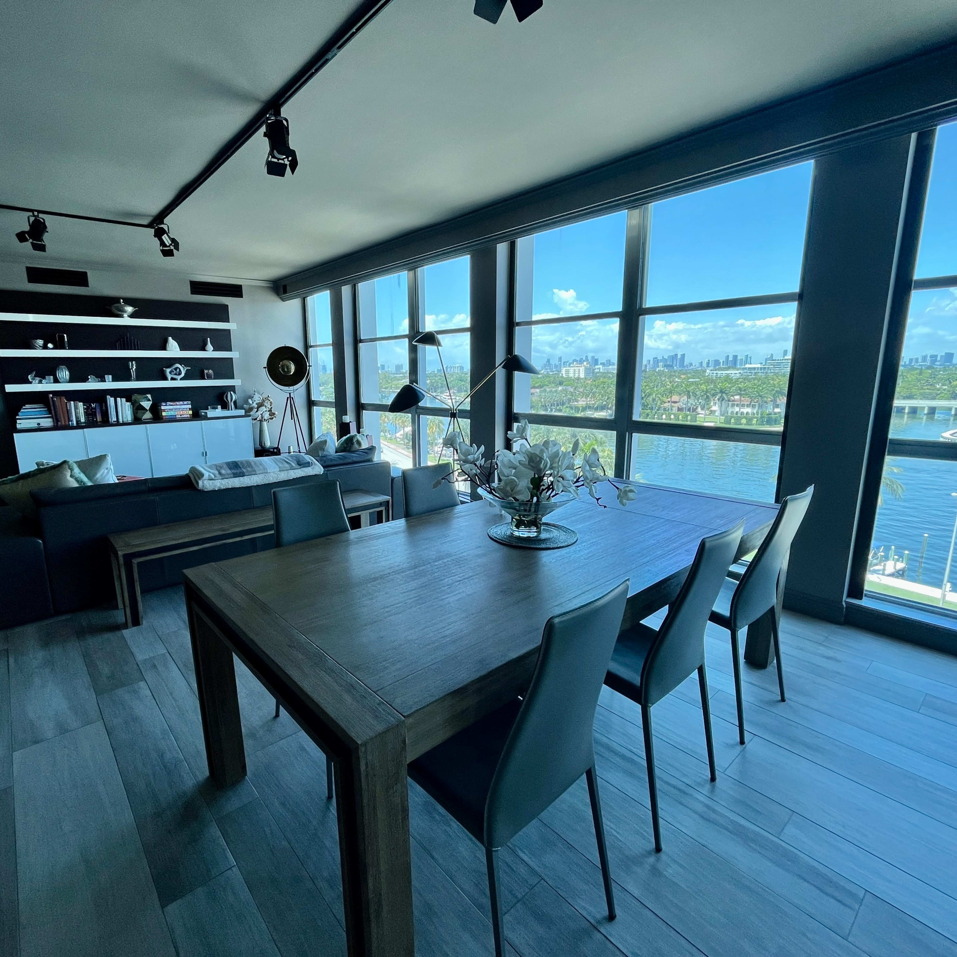 A modern living space featuring a dining table with chairs, a large window overlooking a waterfront view, and shelves filled with decorative items.