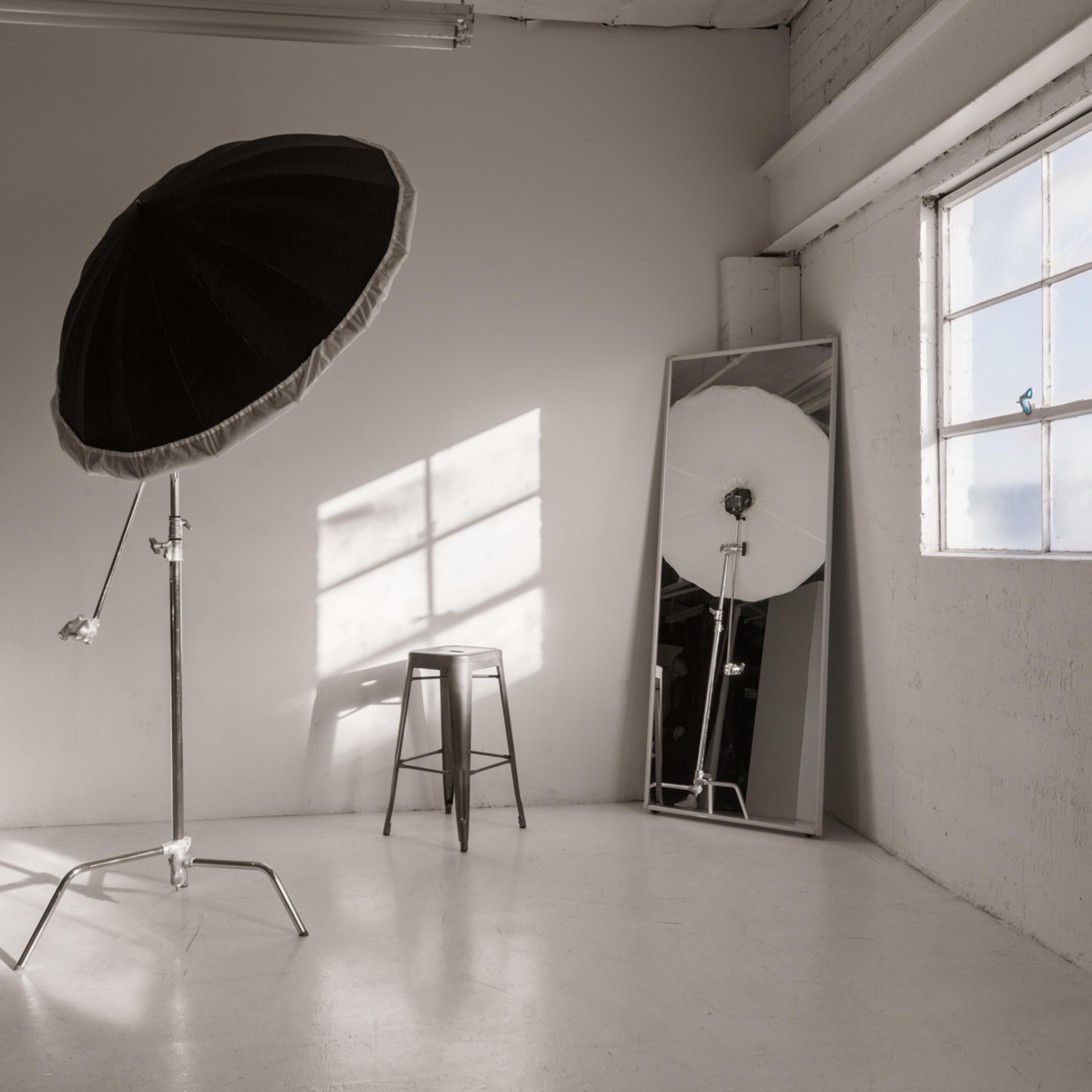 A photography studio features a large softbox, a mirrored wall, a stool, and sunlight casting shadows on the floor.