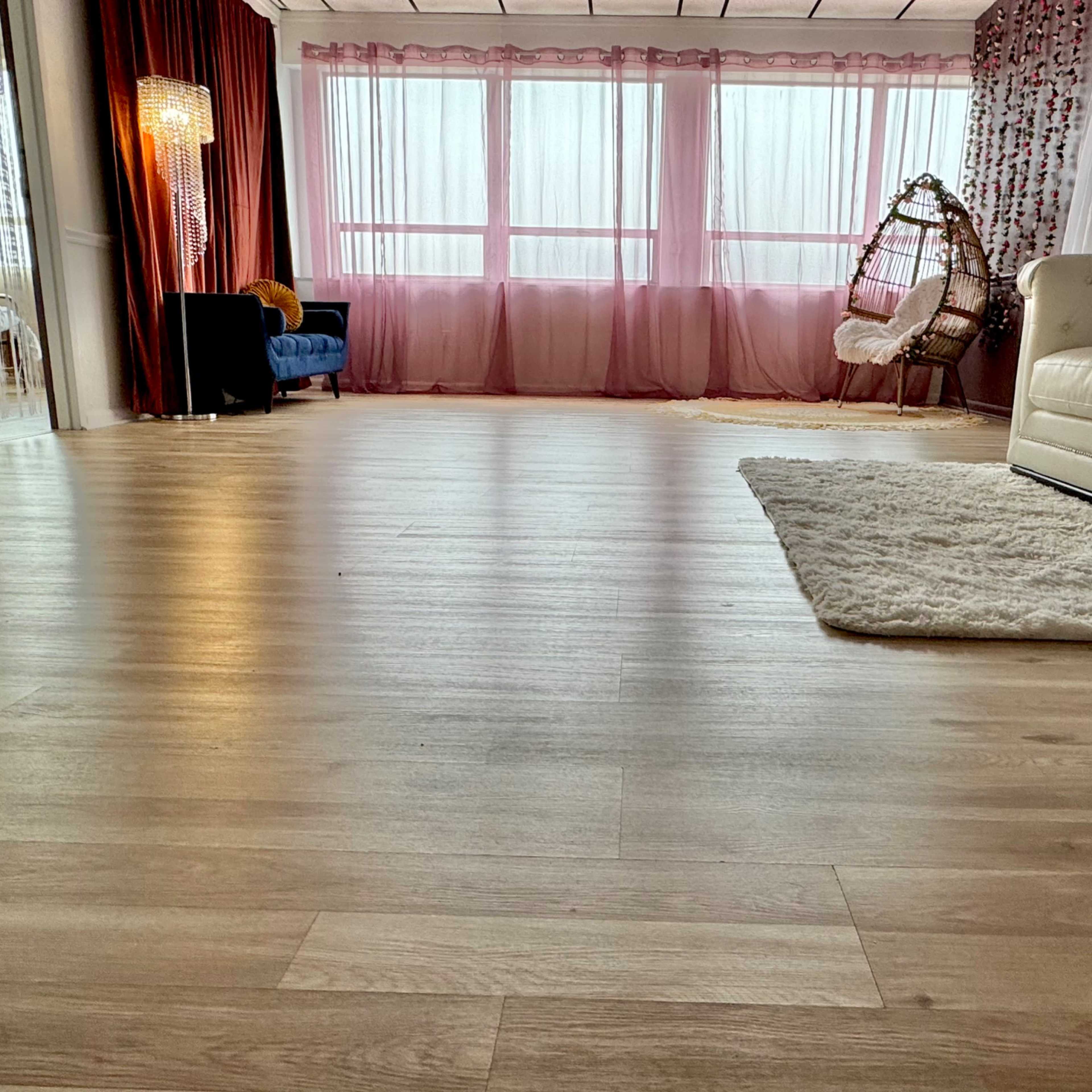 A spacious room with large windows, a wooden floor, a white sofa, a small blue chair, and a hanging chair surrounded by sheer curtains.