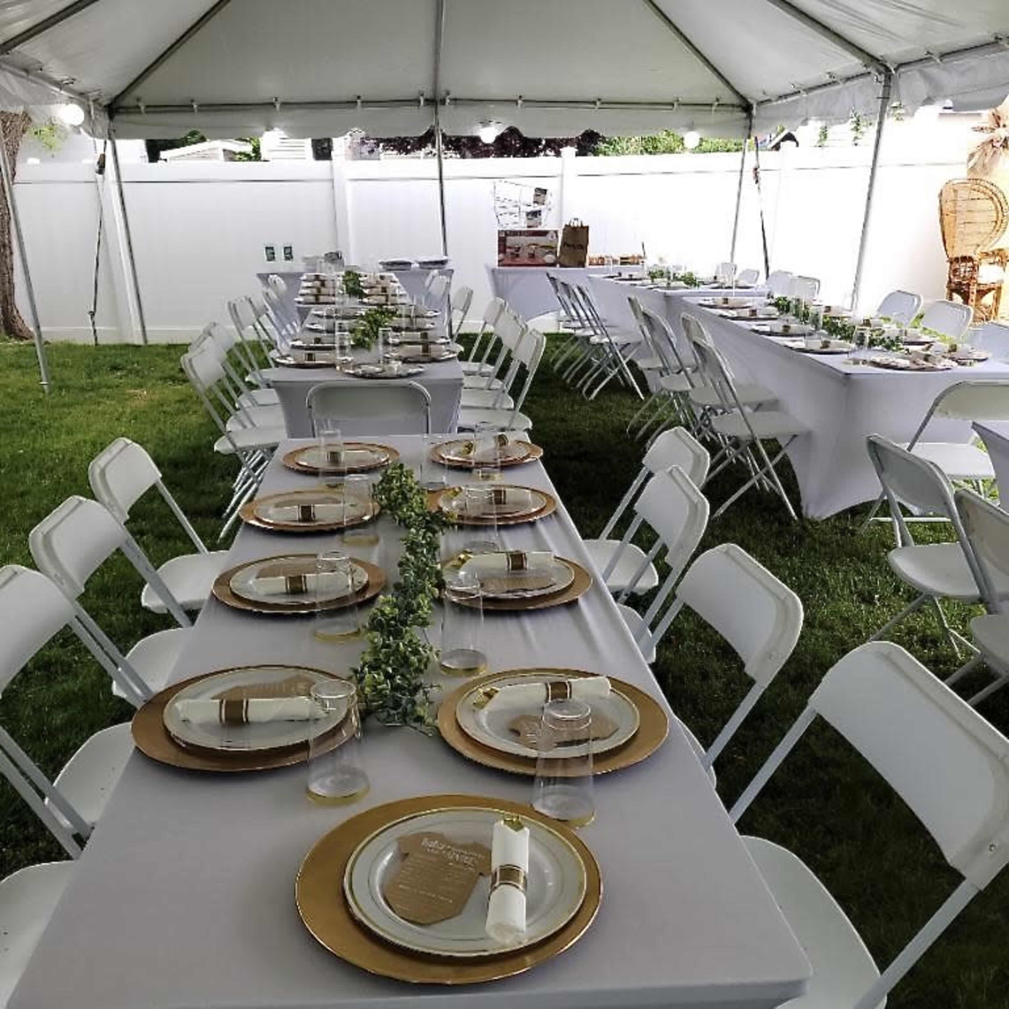 A tented outdoor space is set up with long tables covered in gray linens, featuring gold-rimmed plates and glassware arranged for a formal gathering.