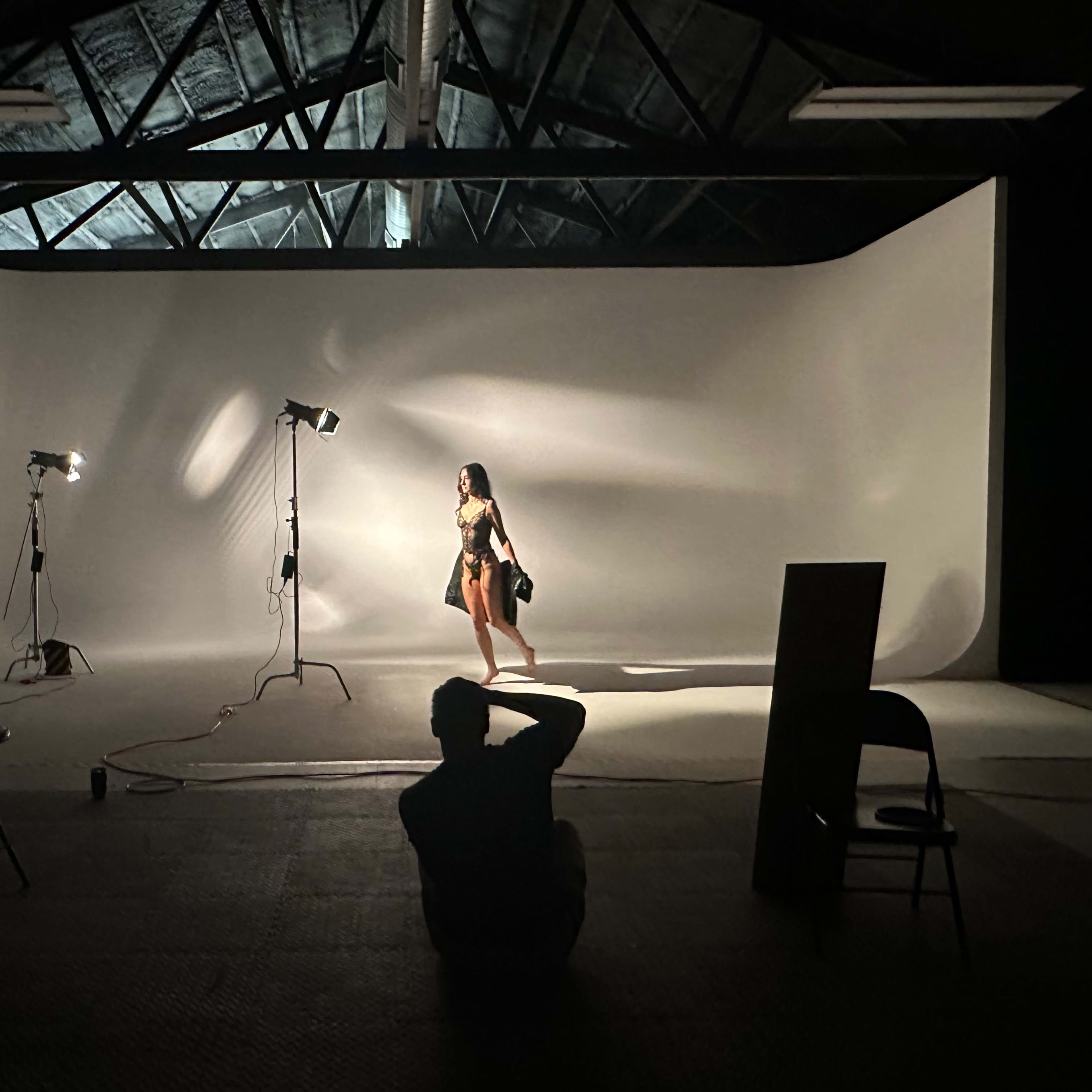 A person poses in a studio with a gray backdrop while a photographer captures the shot from a distance.