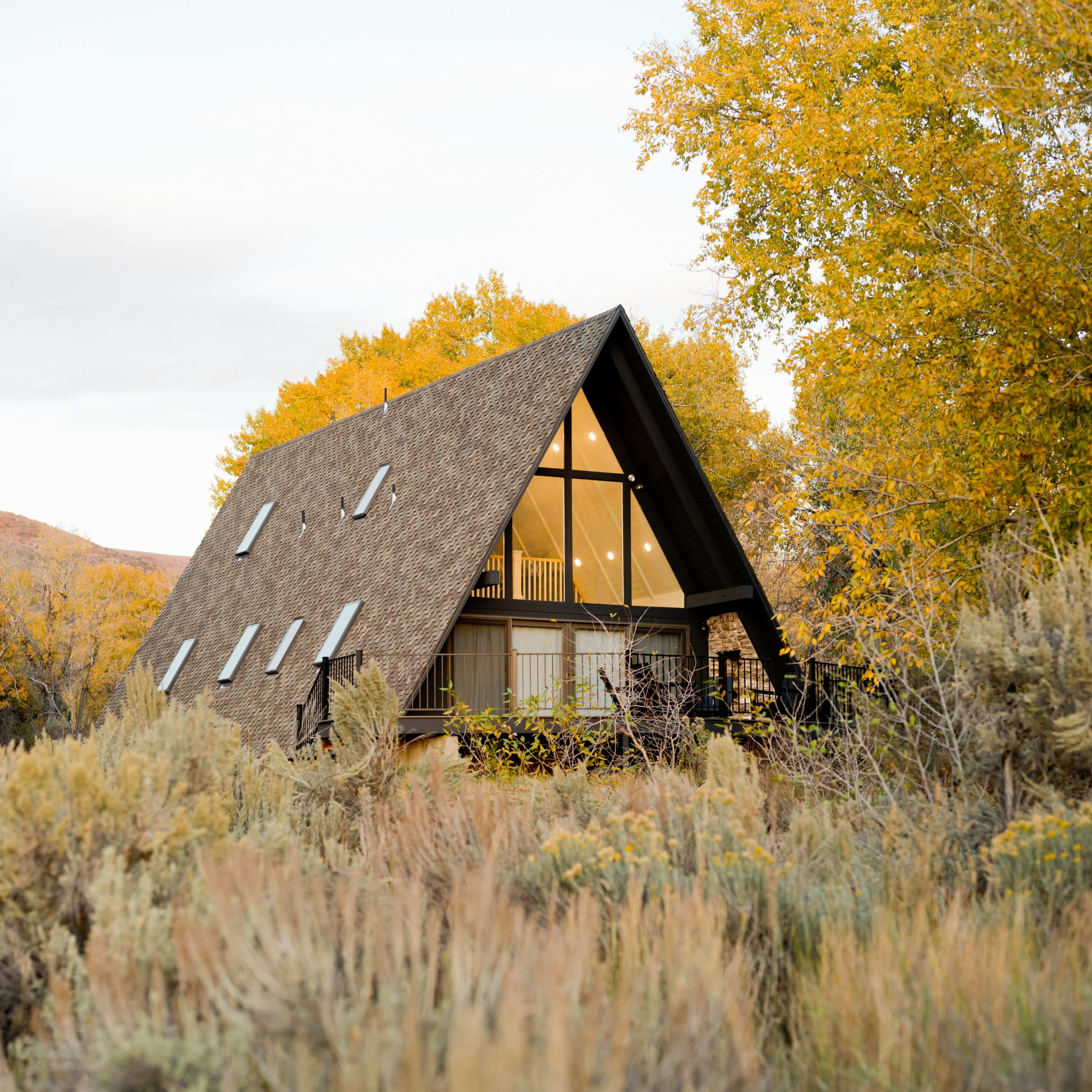 A triangular-roofed cabin is nestled among yellow-leaved trees and shrubs in a rural landscape.