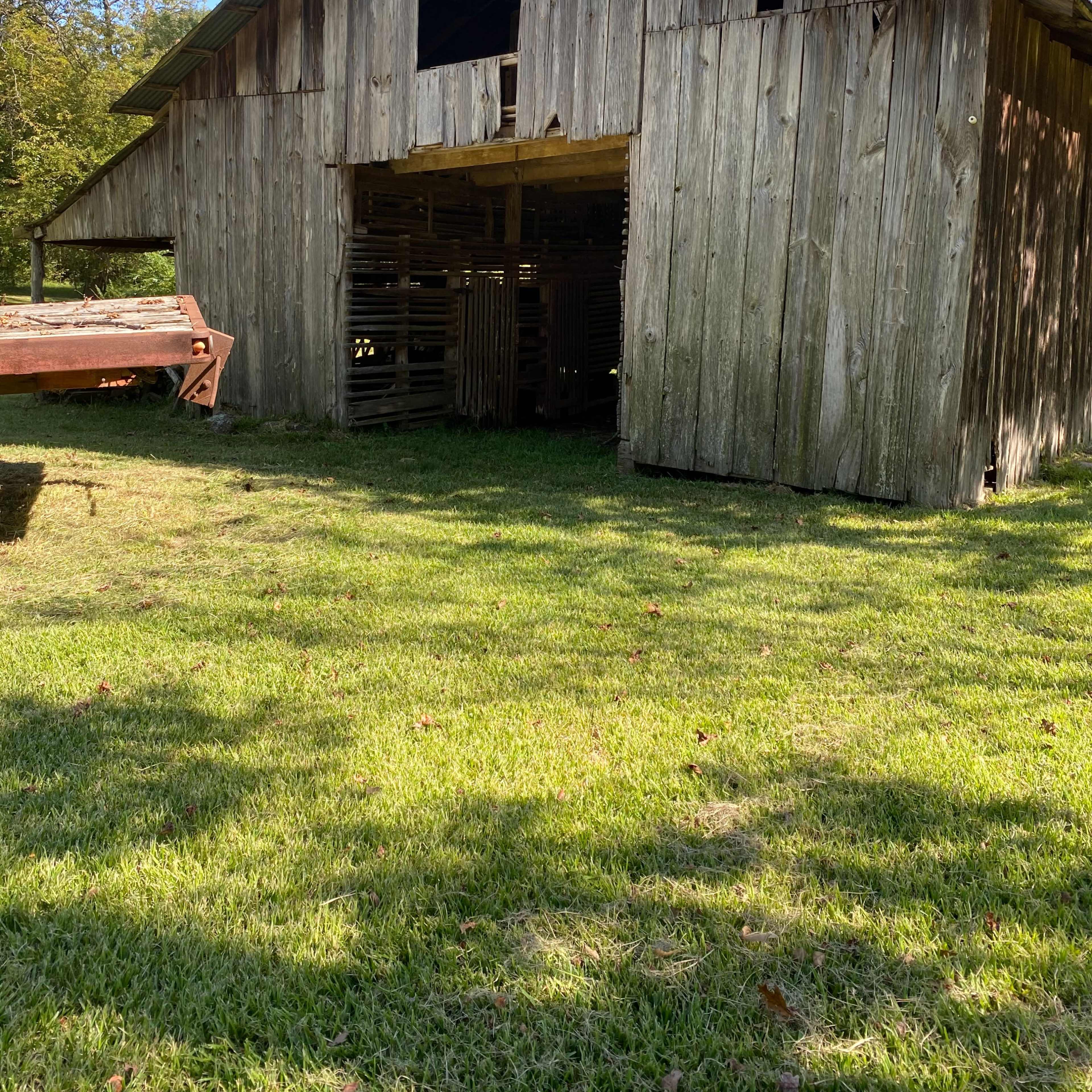 A weathered wooden barn with an open front, situated in a grassy area surrounded by trees.