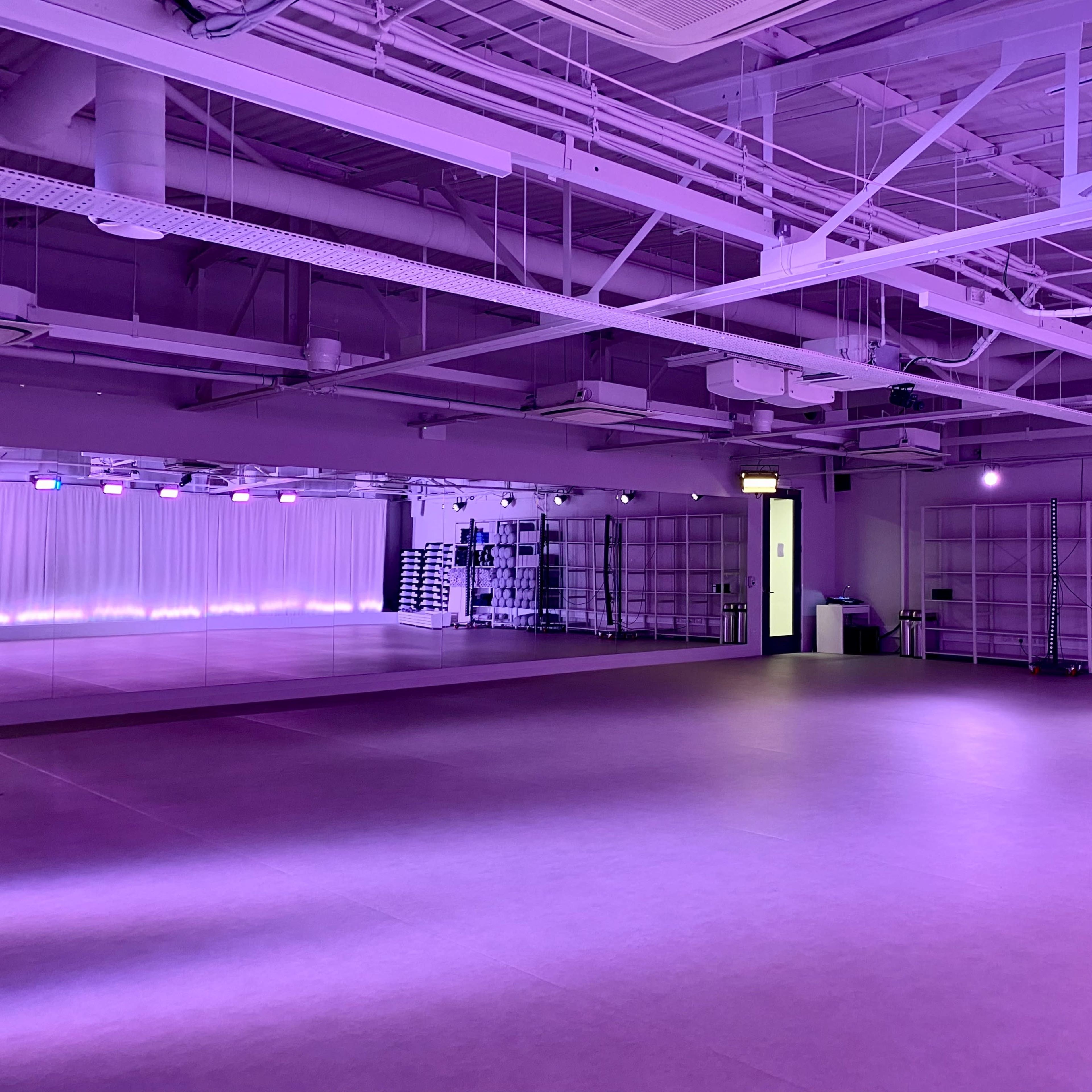 A spacious, well-lit studio with purple lighting, mirrors, and organized shelves holding exercise mats.