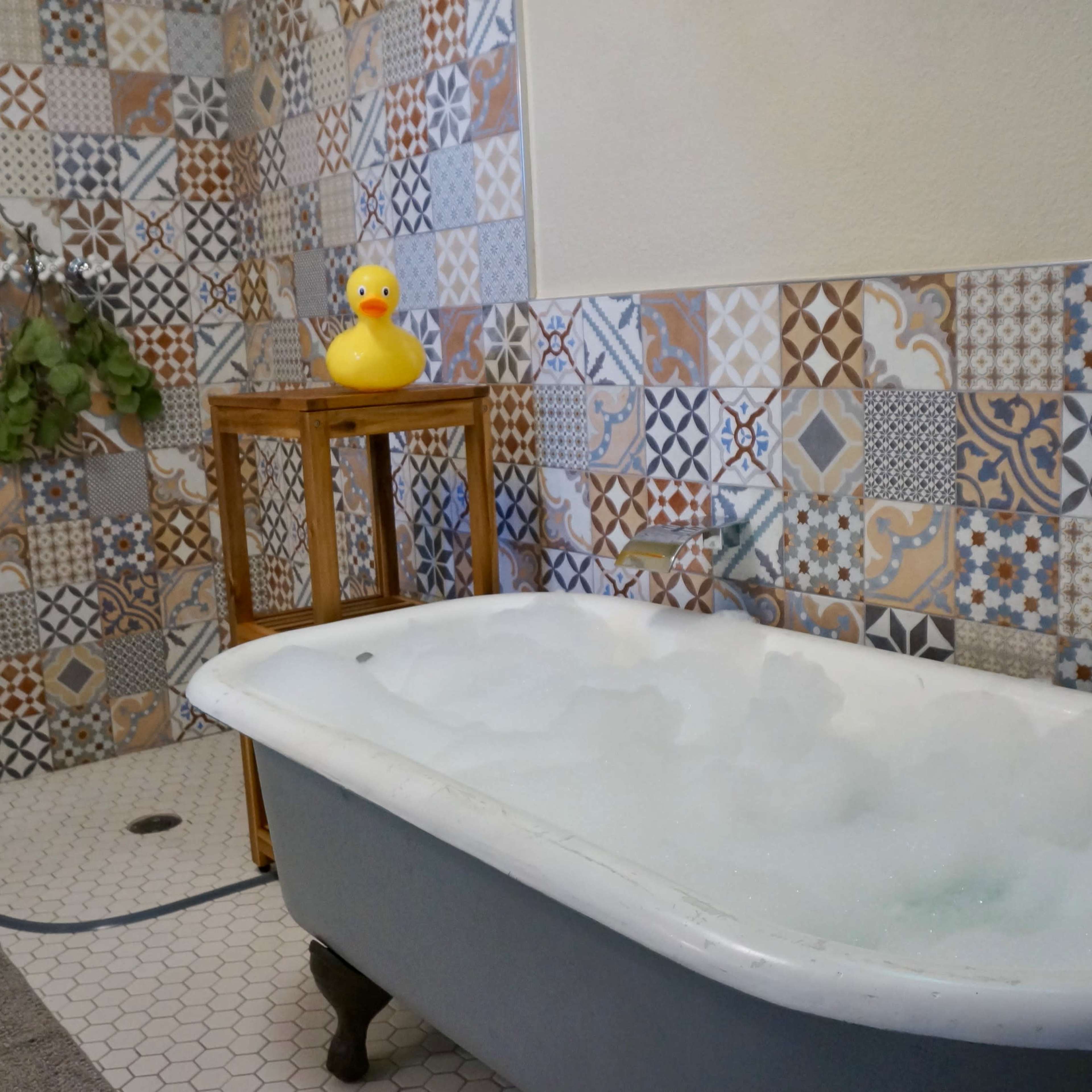 A vintage bathtub filled with bubbles, set in a bathroom with colorful patterned tiles and a wooden side table displaying a rubber duck.